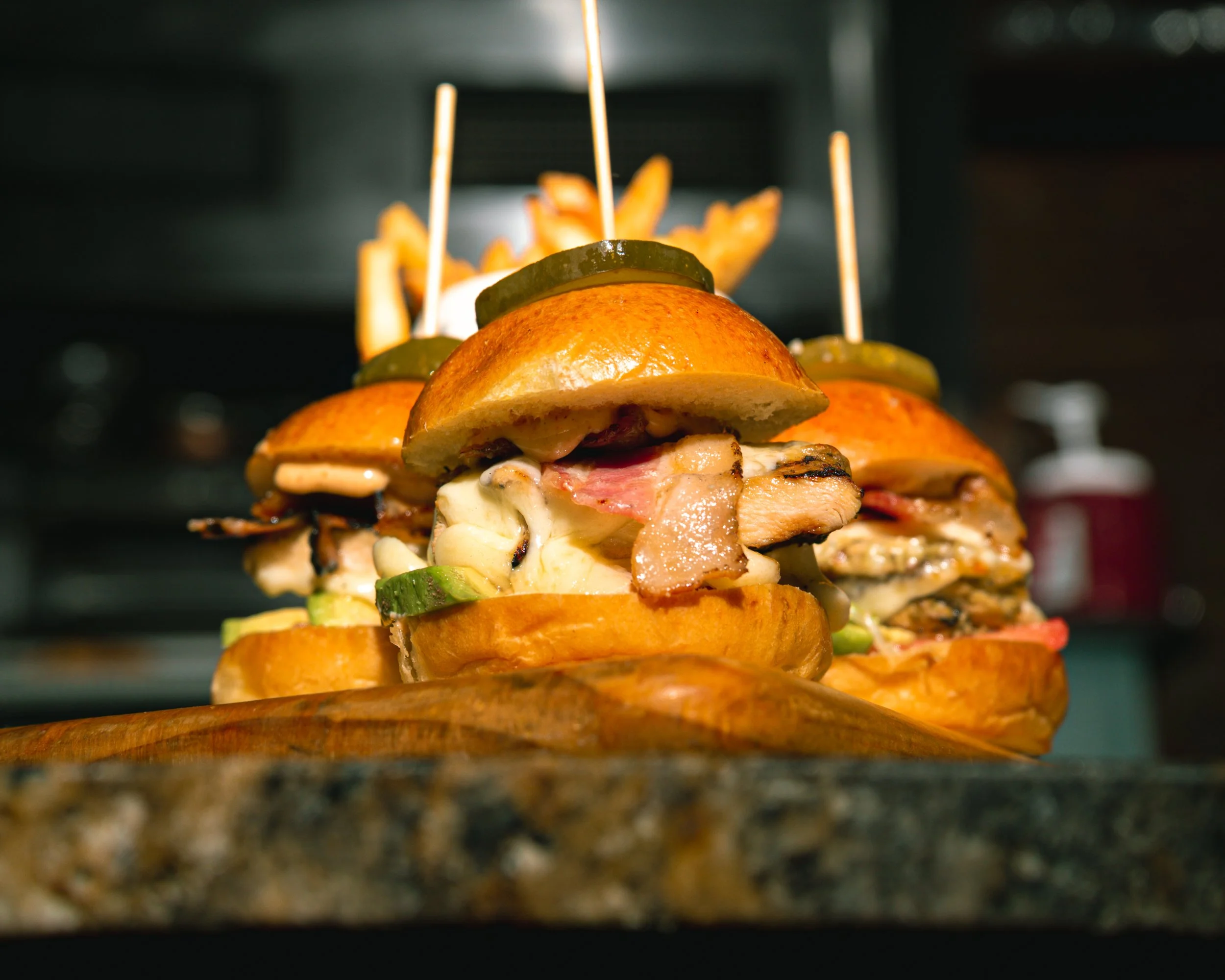 Close-up of stacked burgers with bacon, pickles, and melted cheese, with french fries in the background.