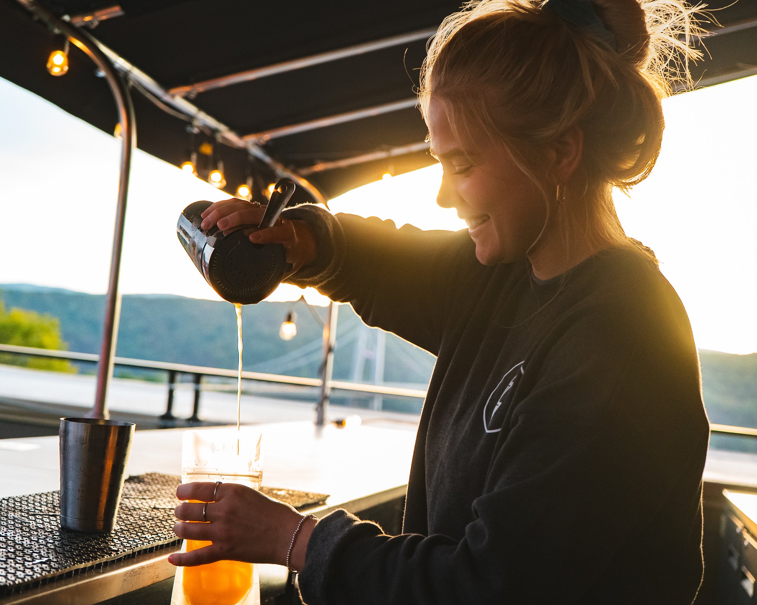 A woman with blonde hair, smiling with eyes closed, holds a glass of orange juice on a table outdoors at sunset.