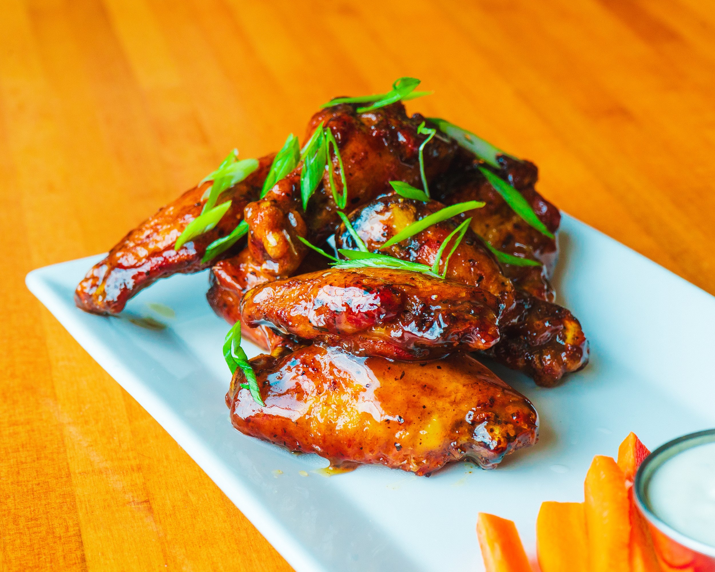 Close-up of glazed chicken drumsticks garnished with green onions served on a white rectangular plate, with sliced vegetables on the side, placed on a wooden surface with an orange cloth.
