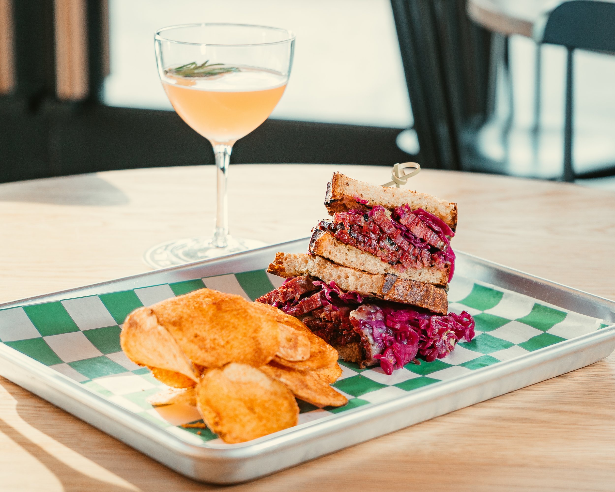 A plate with toasted sandwich halves filled with pinkish-red meat and purple shredded cabbage, along with potato chips, on a patterned paper tray, with a glass of pink drink garnished with a sprig of rosemary in the background.