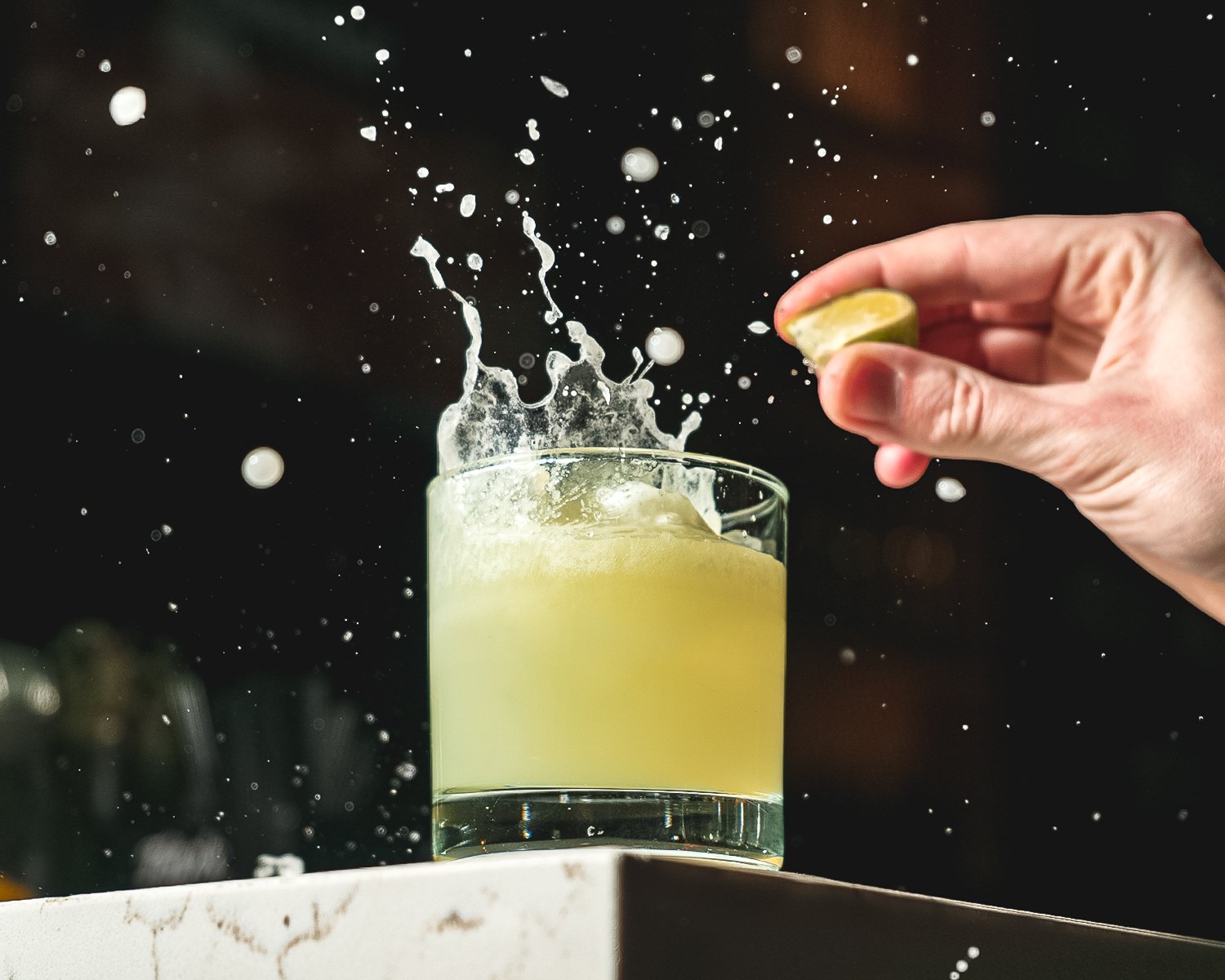 A hand squeezing a lime into a glass of lemonade, causing splashes.