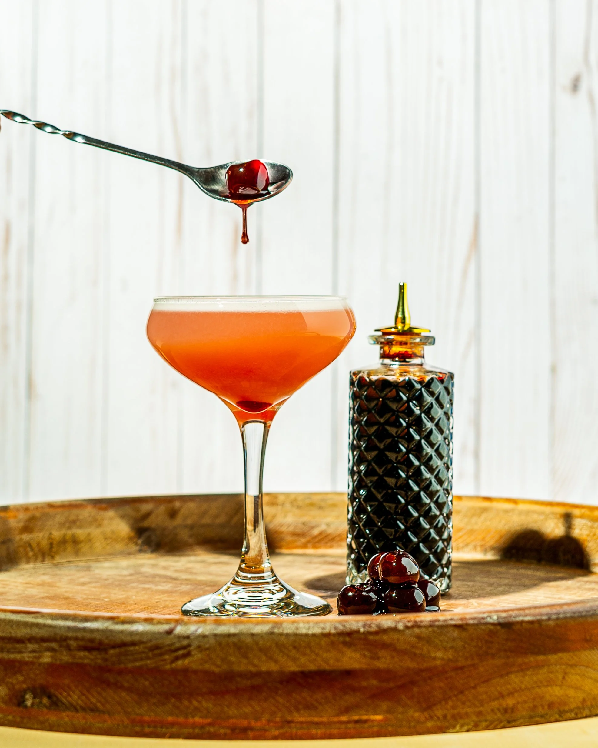 A cocktail glass with an orange-colored drink, a spoon with a cherry on it above the glass, a bottle with a dropper, and cherries on a wooden surface, against a wooden wall background.