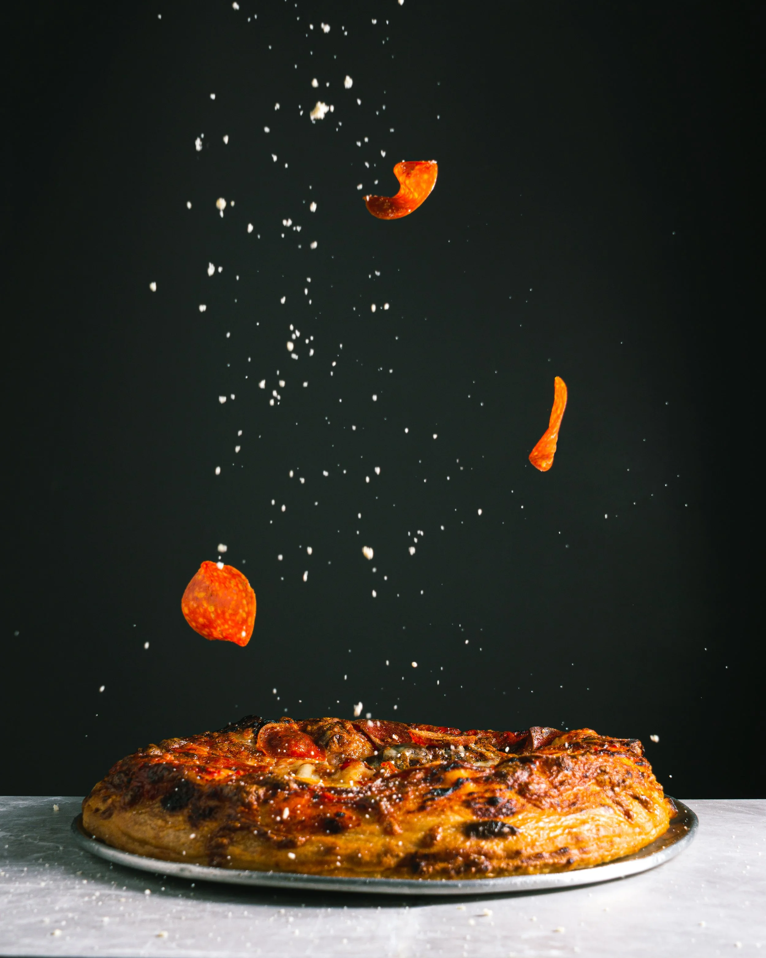 A large, cooked pepperoni pizza on a plate with pepperoni slices falling onto it from above against a black background.