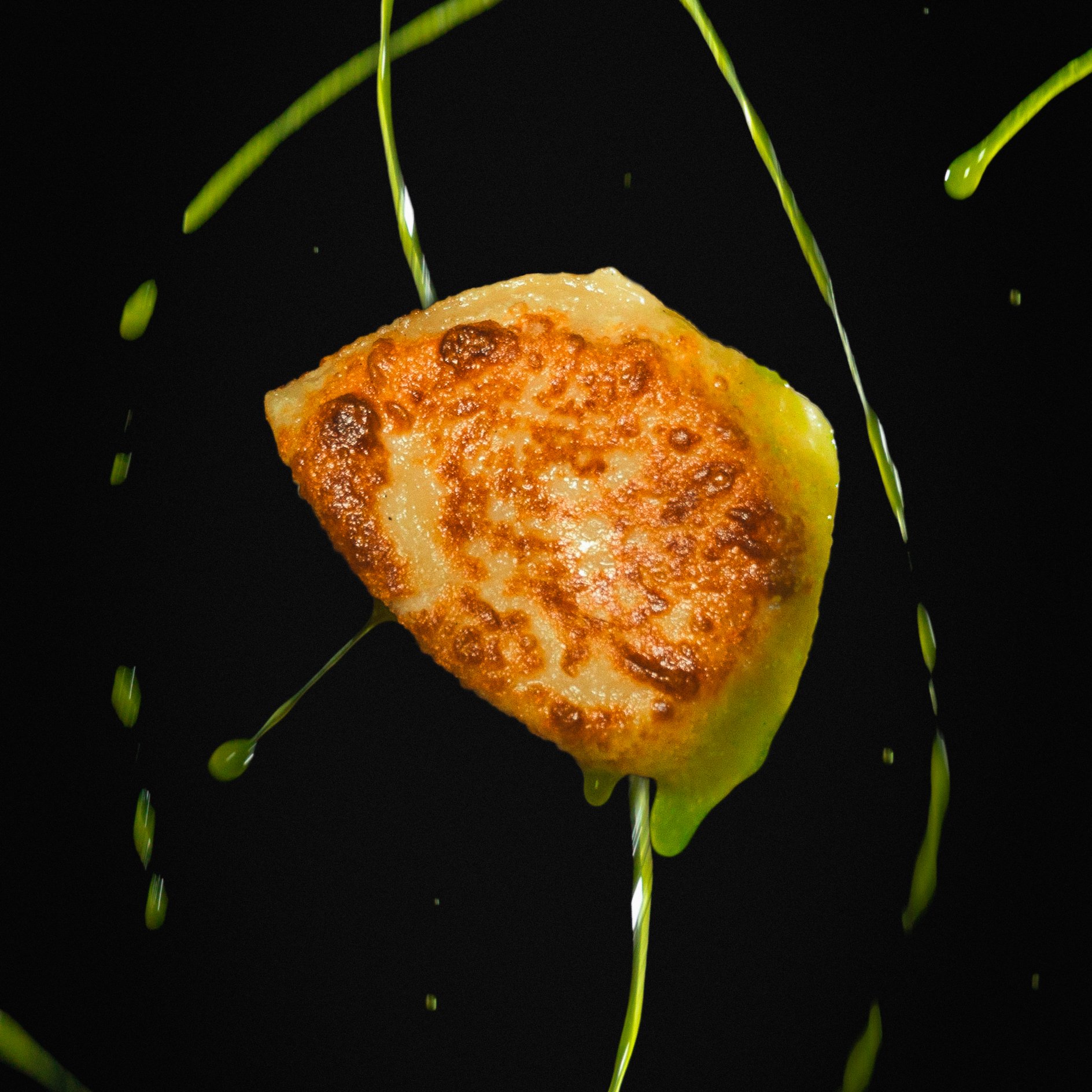 Close-up of a fried cheese ball with melted cheese oozing out, surrounded by green sprouts and drops of green oil against a black background.