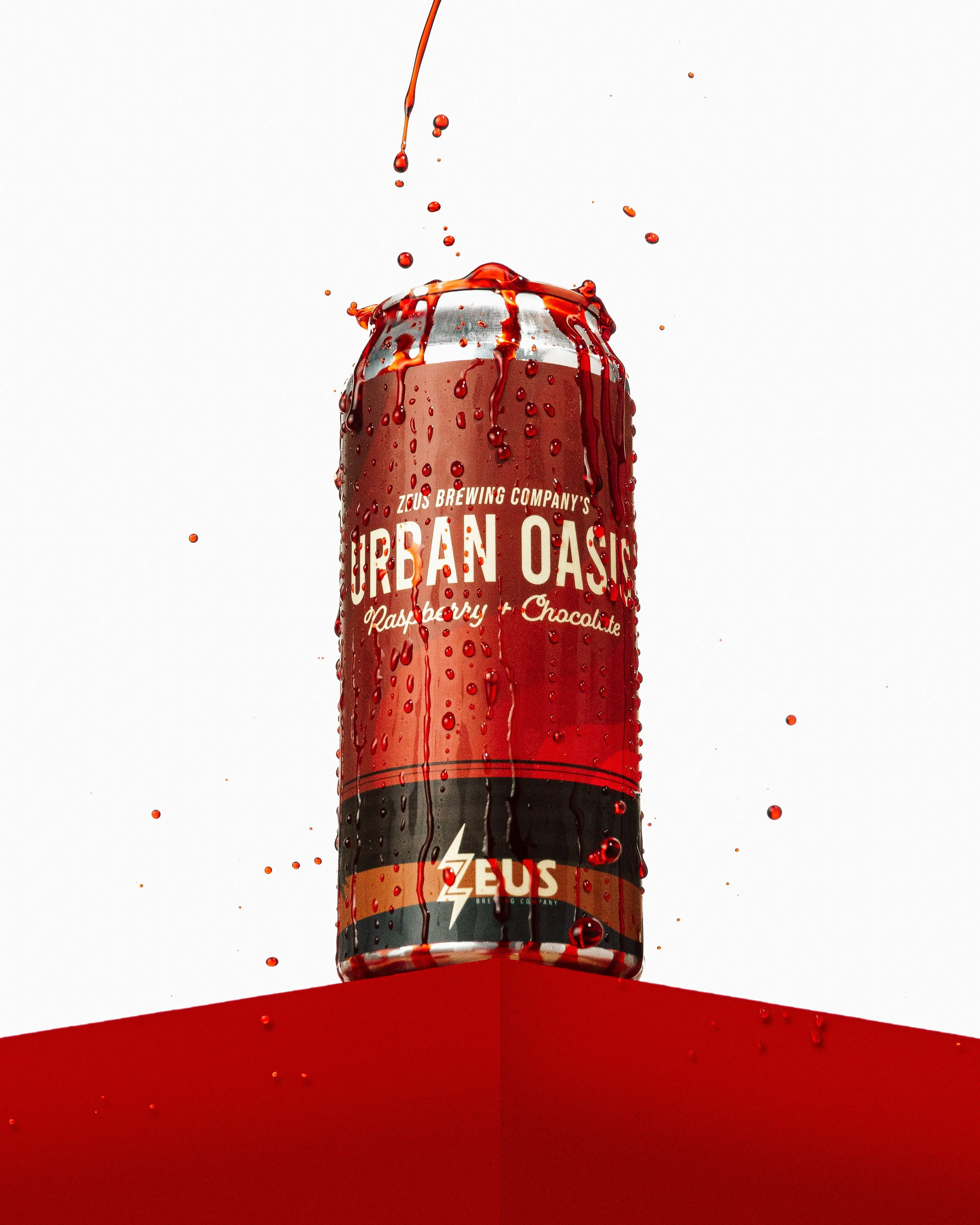 A can of Zeus Brewing Company's 'Urban Oasis' Raspberry Chocolate beverage with red liquid splashing over the top, droplets scattered around, against a white background.