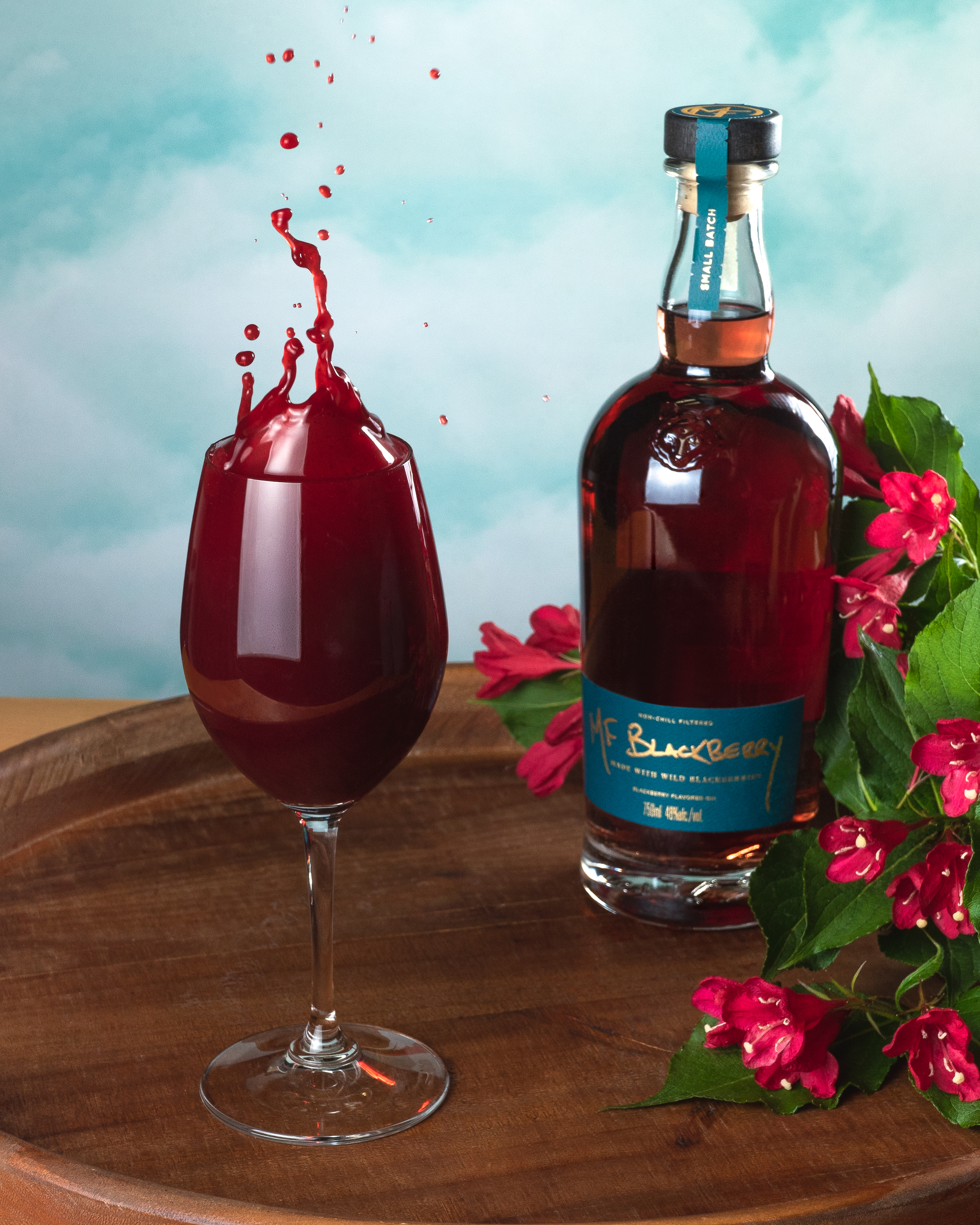 A wine glass with red liquid splashing inside next to a bottle of blueberry flavored beverage and pink flowers on a wooden tray with a cloudy blue sky background.