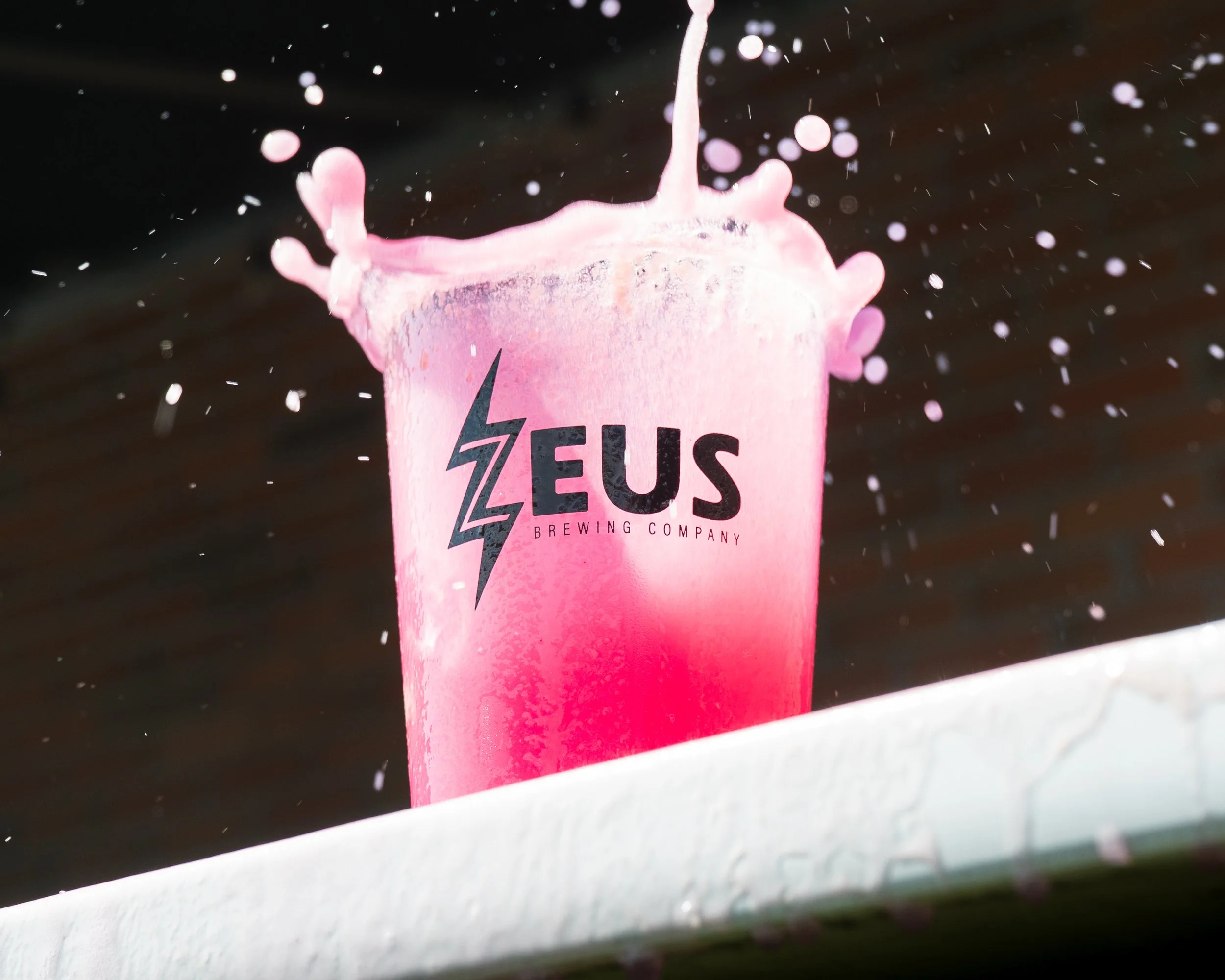 Pink cup from Zeus Brewing Company with liquid splashing out as viewed through a vehicle windshield with raindrops
