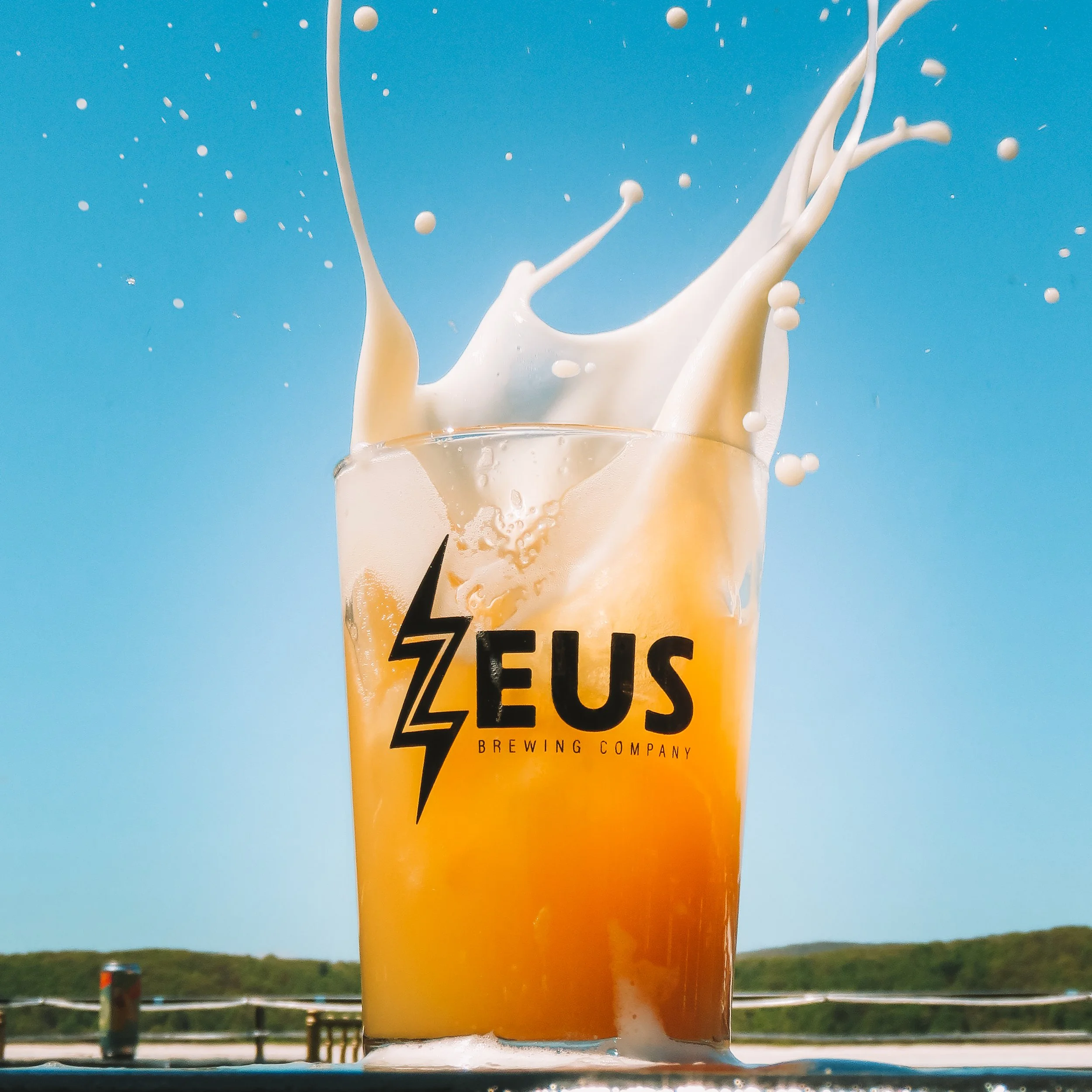 A glass of orange-colored beverage with a splash of cream or milk erupting from the top, set against a blue sky background.