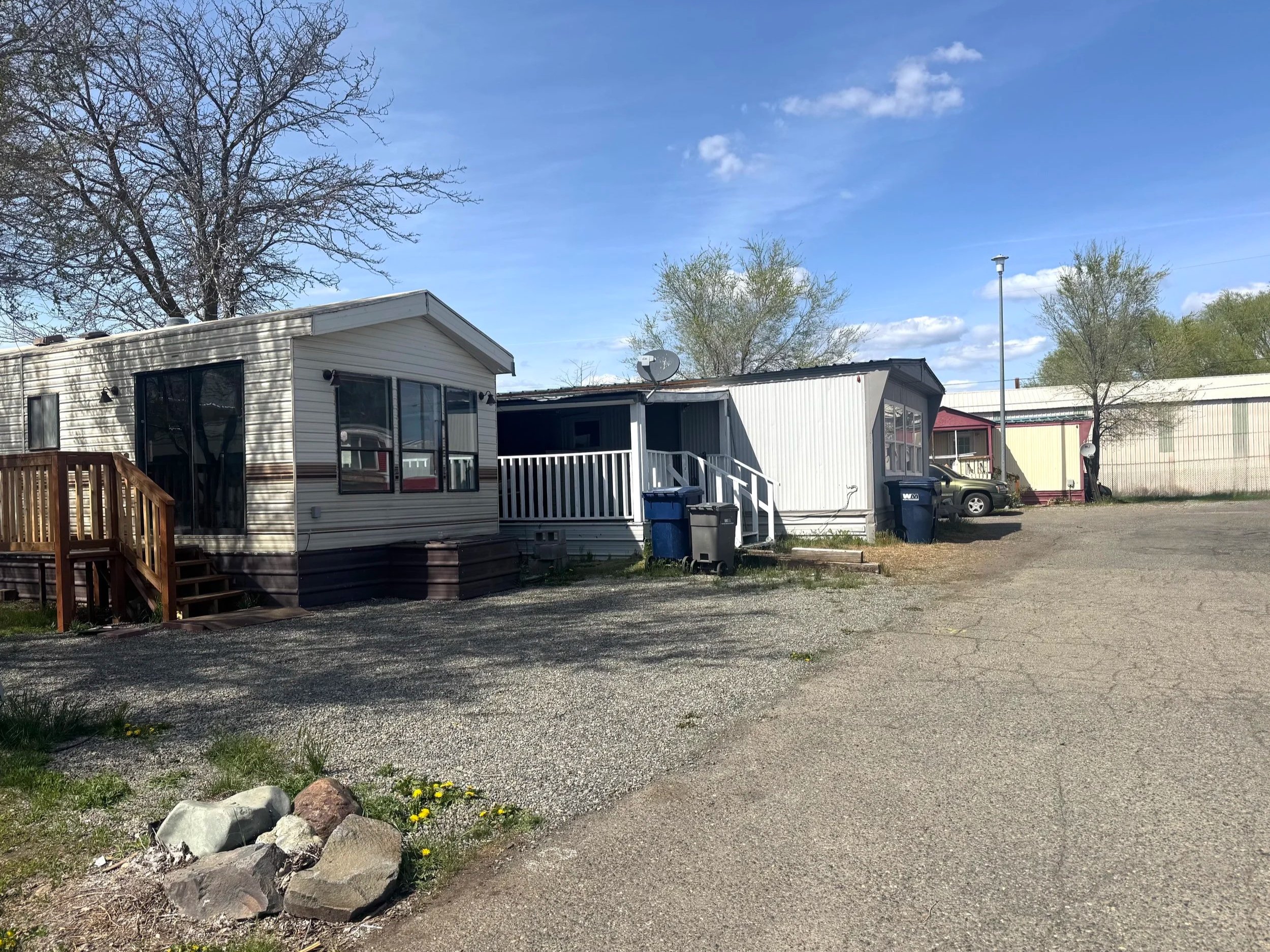 Affordable mobile homes and trailers in a rural area with gravel parking lot, trees, and blue sky.