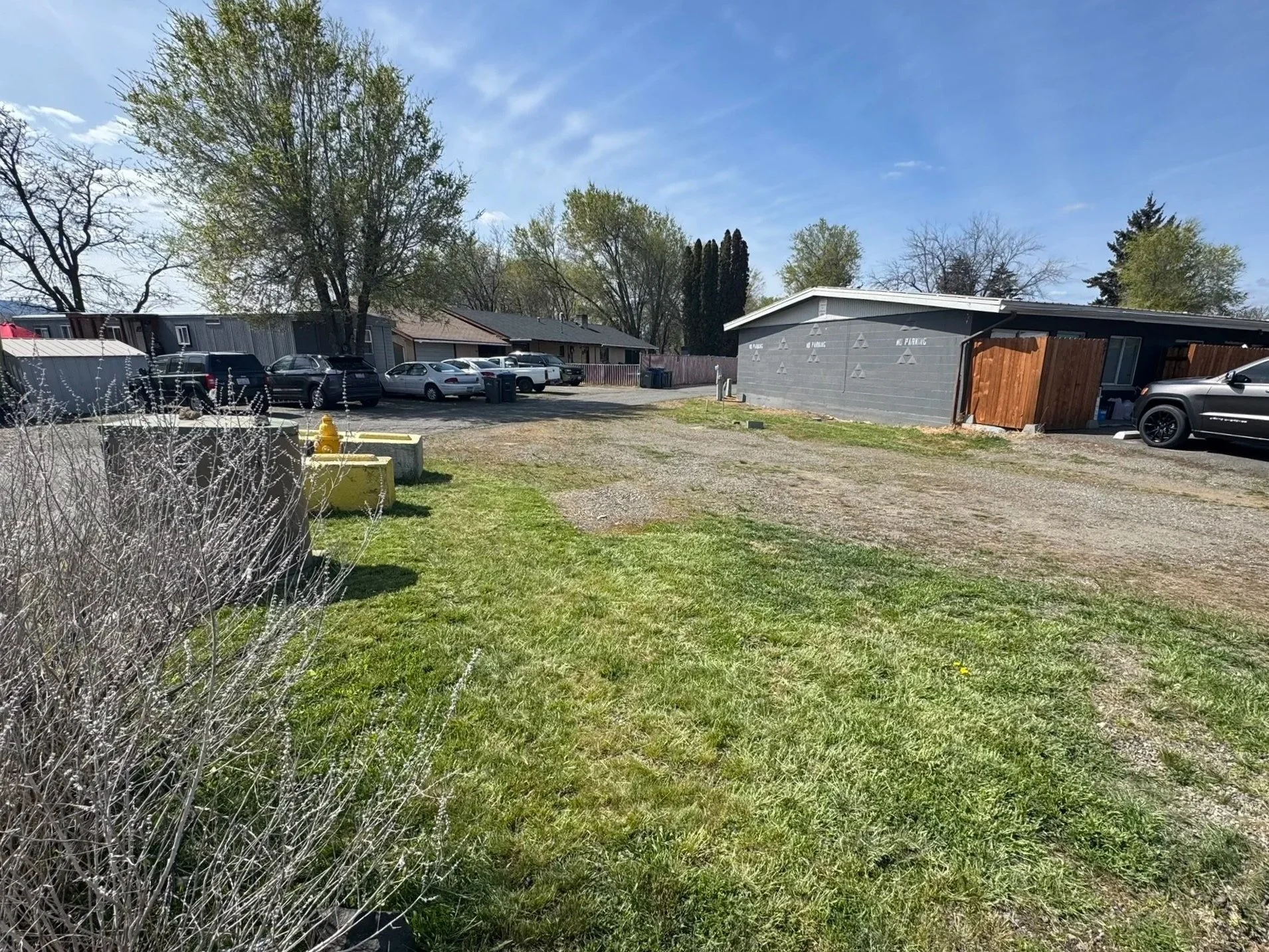 A parking lot with several parked cars, a grassy area with bushes in the foreground, trees and residential houses in the background, and a building painted gray with a wooden fence on one side under a partly cloudy sky.