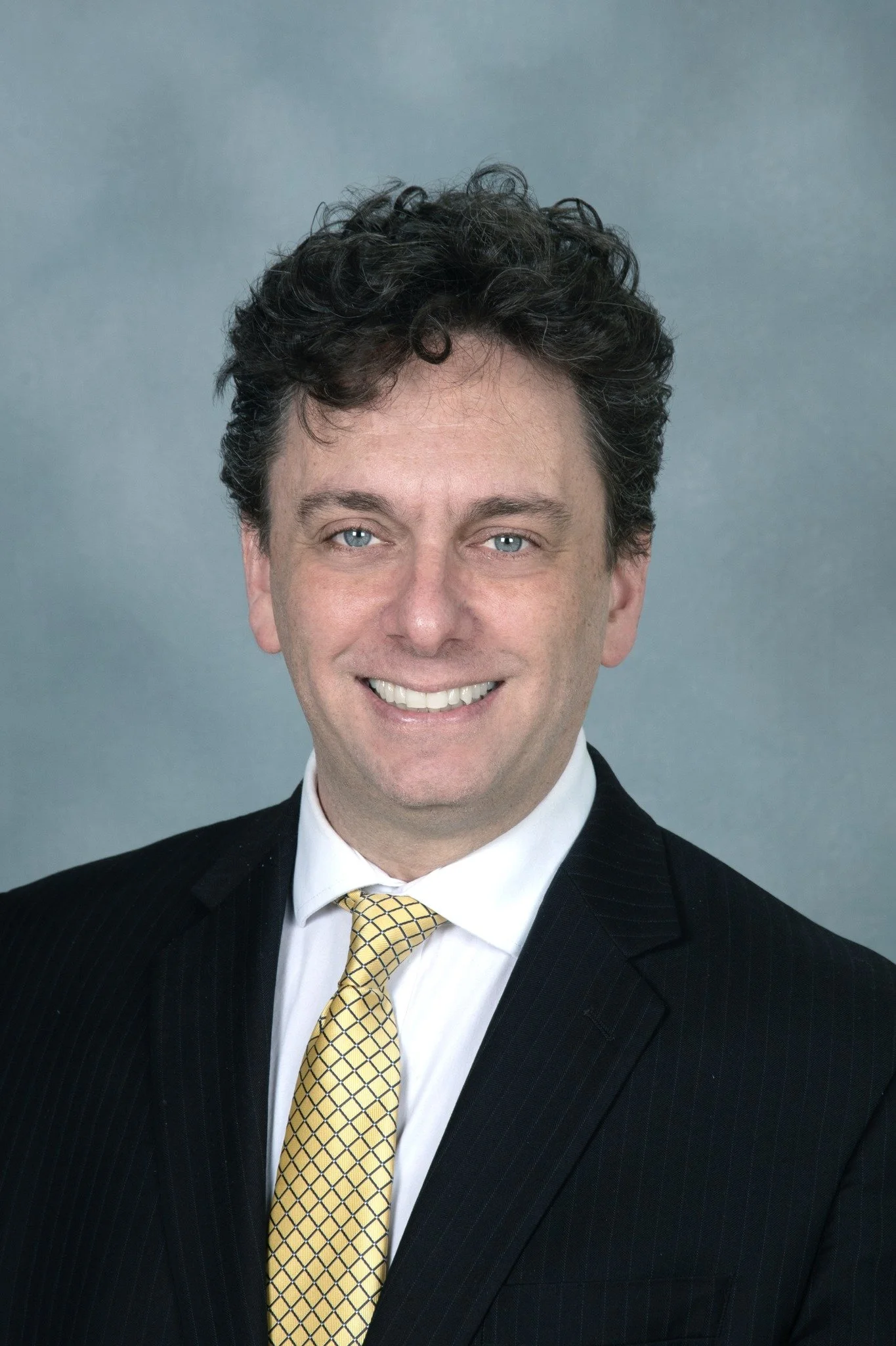 A man in a black suit with a white shirt and yellow checkered tie, smiling, with short, curly dark hair, against a plain gray background.