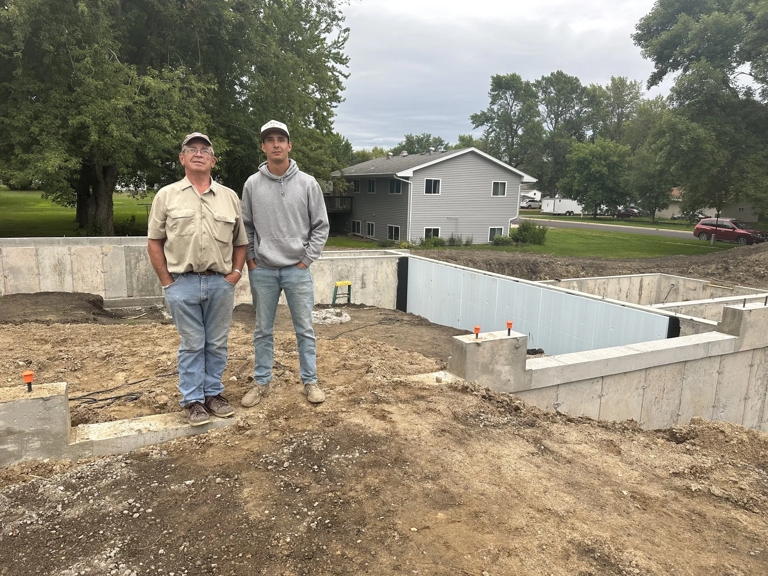 Two men standing side by side on a construction site with a house foundation under construction behind them, in a suburban area with trees and houses in the background.