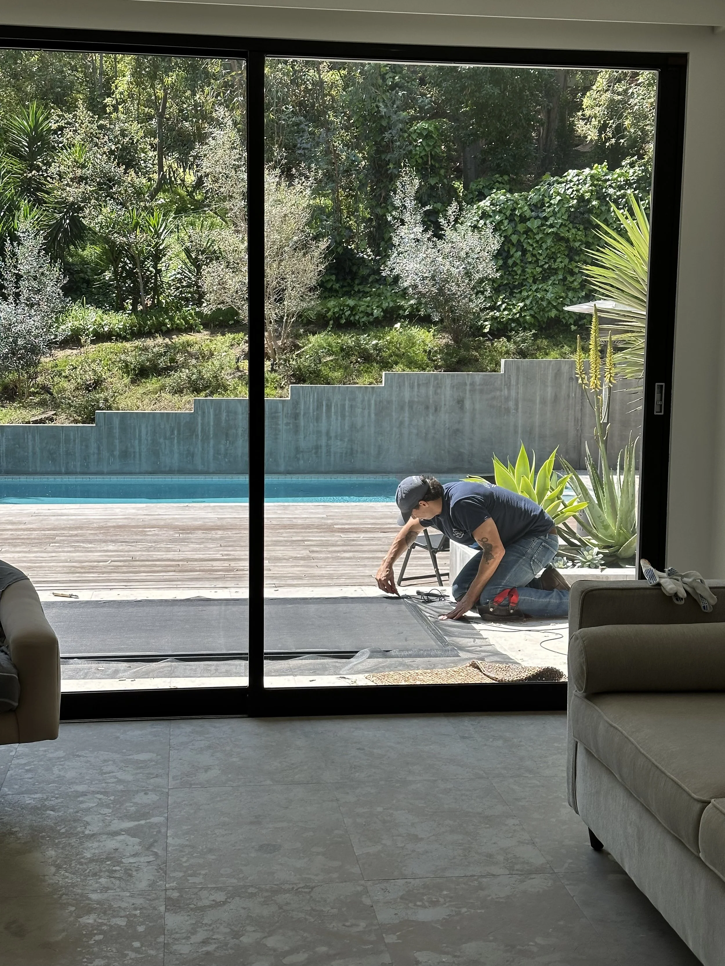 A man is kneeling outside on a wooden deck, working on the sliding glass door, with lush green bushes and trees in the background.