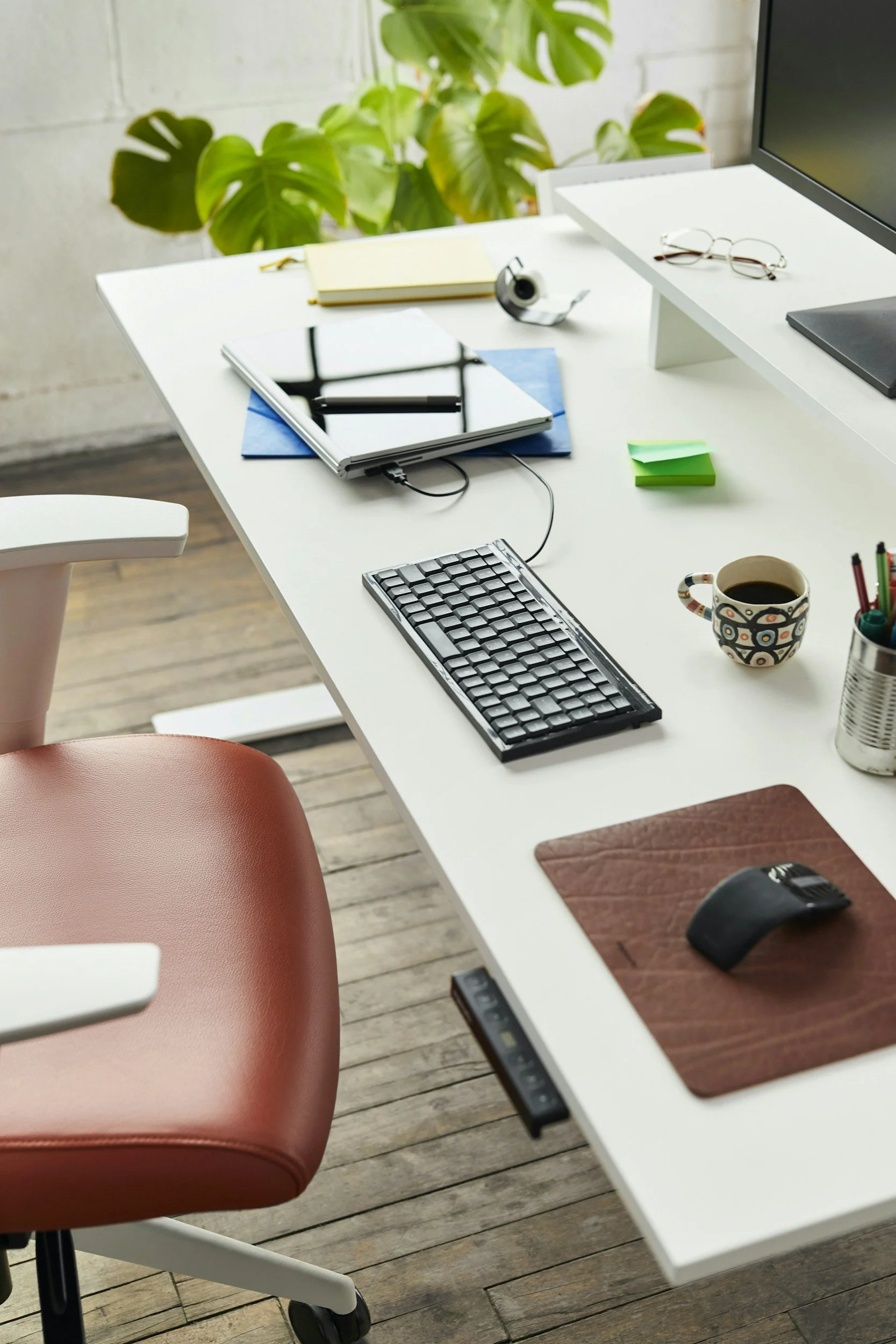 An organized white desk with a computer monitor, keyboard, mouse on a wooden desk mat, and a cup of coffee. Items include a closed laptop, glasses, sticky notes, a tape dispenser, a notepad, a tablet, and a container with pens and pencils. A plant with large green leaves is visible in the background.