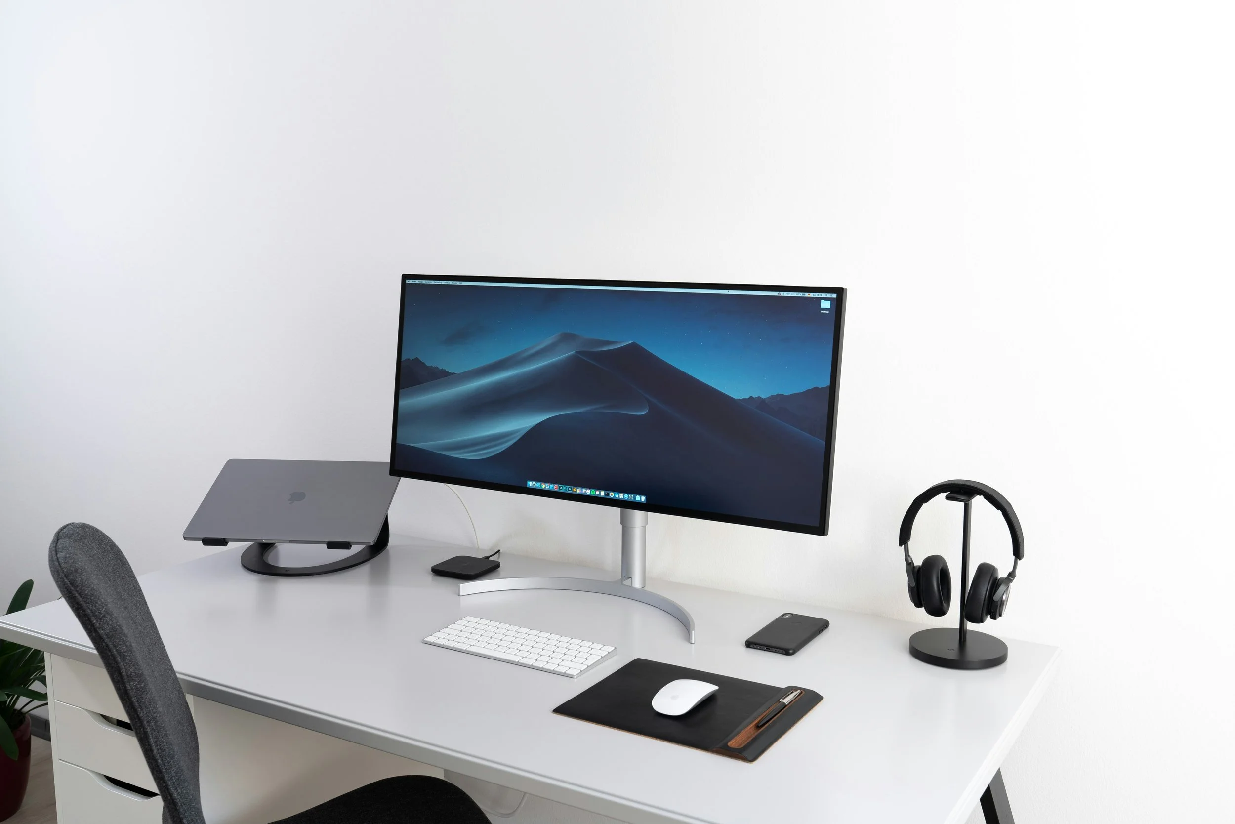 Modern minimalist workspace with a large curved monitor, a wireless keyboard and mouse on a black desk mat, a closed laptop on a stand, a smartphone, and a pair of black headphones on a stand, all on a white desk against a plain white wall.
