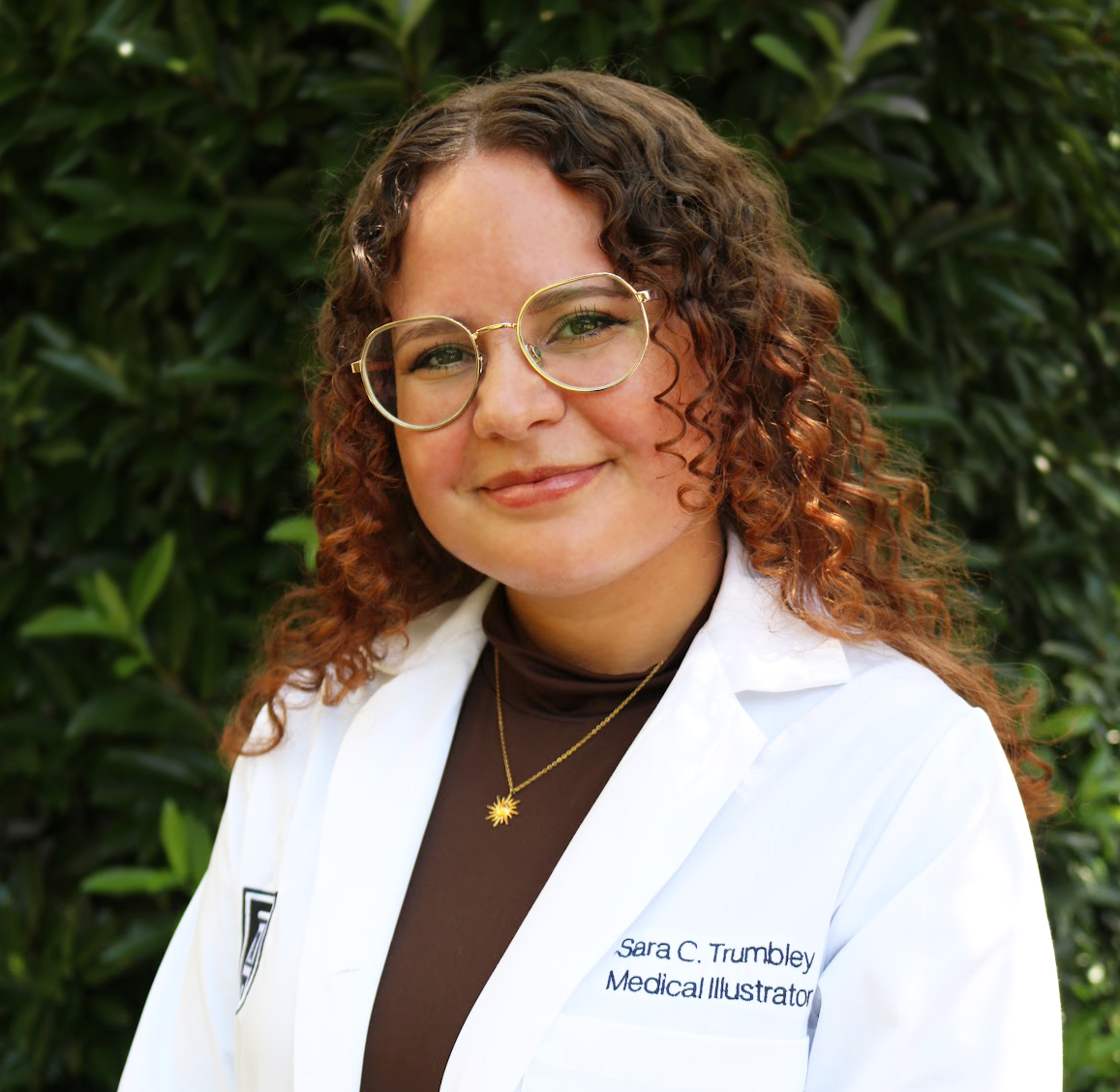 Portrait of a woman with curly brown hair and glasses, wearing a white medical coat with her name and title embroidered, standing in front of green foliage.