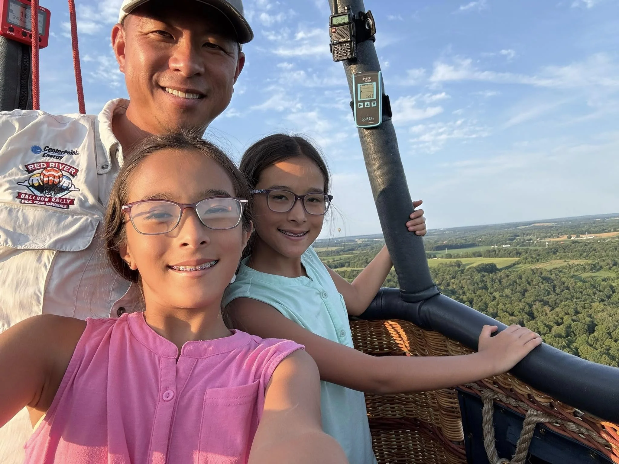 Pilot Ben and his two daughters from the hot air balloon basket, smiling for a selfie with a scenic landscape of greenery and distant fields under a partly cloudy sky.