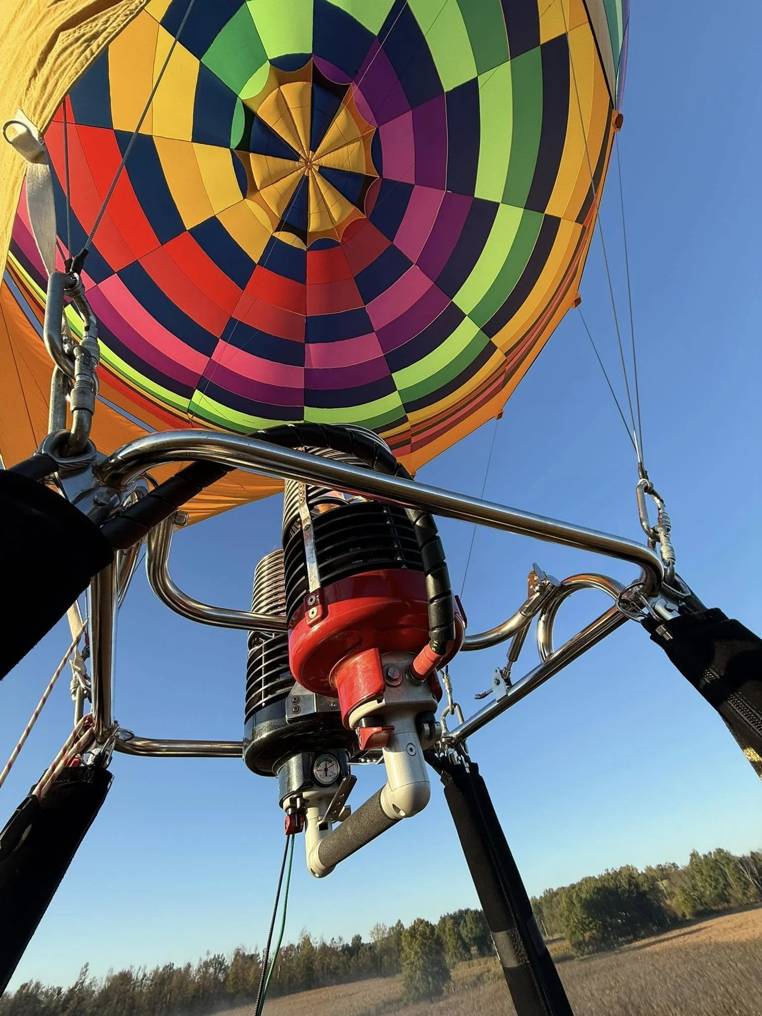 Inside a hot air balloon basket, looking up toward the colorful balloon envelope, with a clear blue sky and trees in the background.