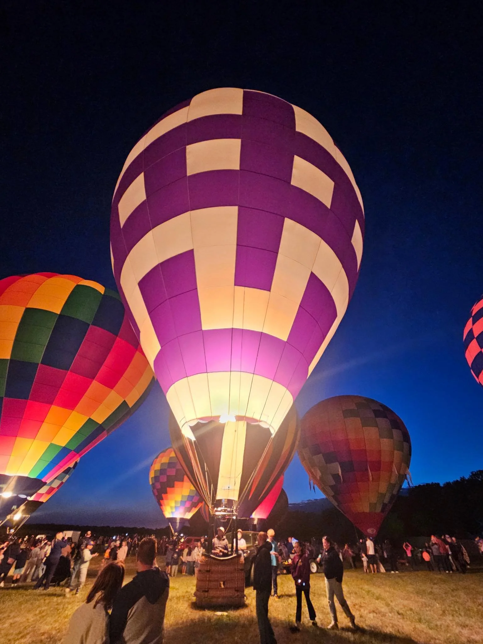 Nighttime scene of hot air balloons illuminated and "glowing", a crowd pleaser while balloons are tethered to the ground.