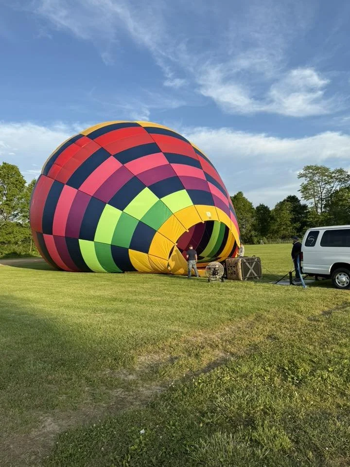 A colorful hot air balloon partially inflated on a grassy field, with people working on it, a white van nearby, and a blue sky with some clouds overhead.