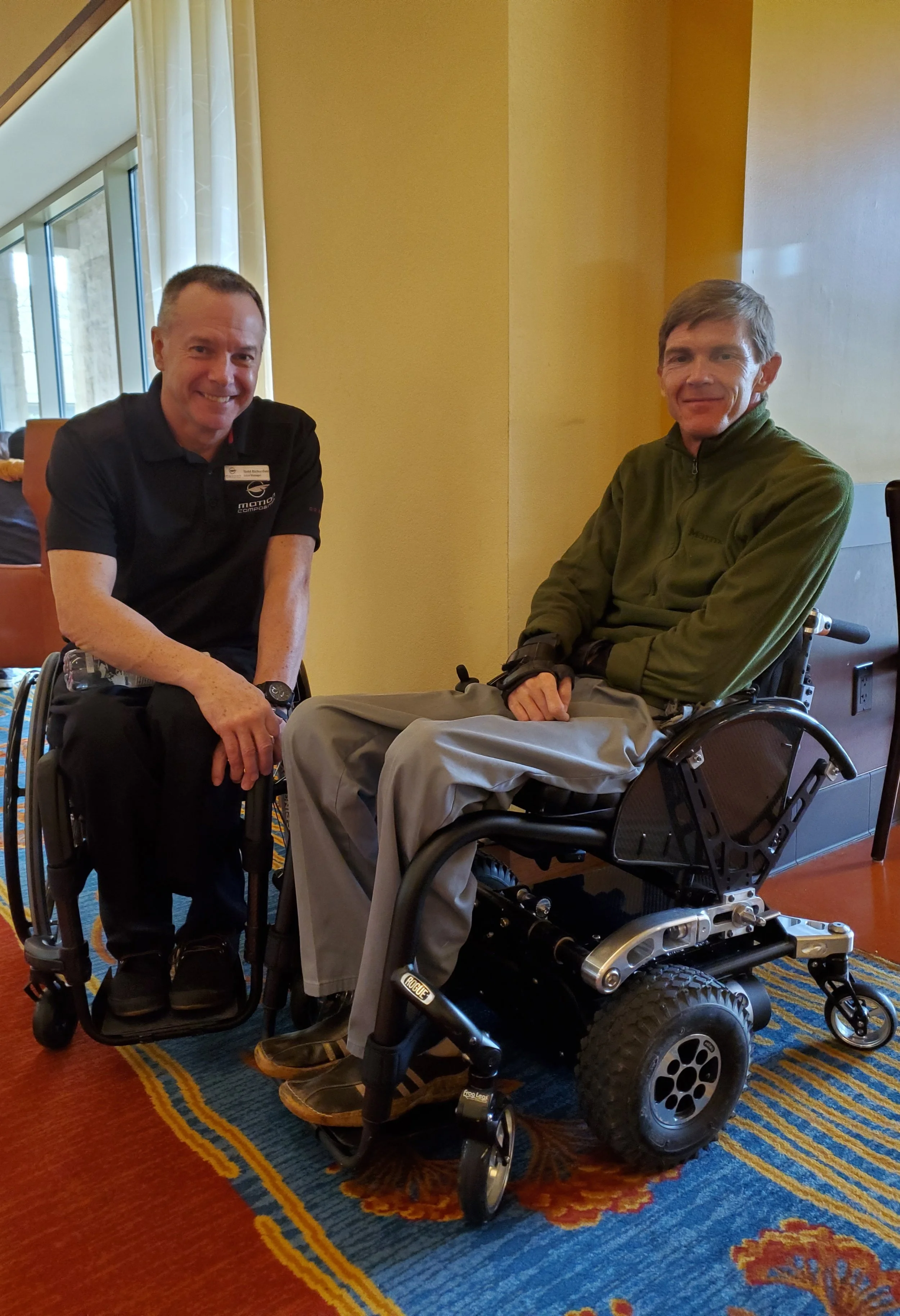 Two men in chairs, one in an electric wheelchair, smiling at the camera in a well-lit room with yellow walls and large windows, carpet with a red, blue, and yellow pattern.