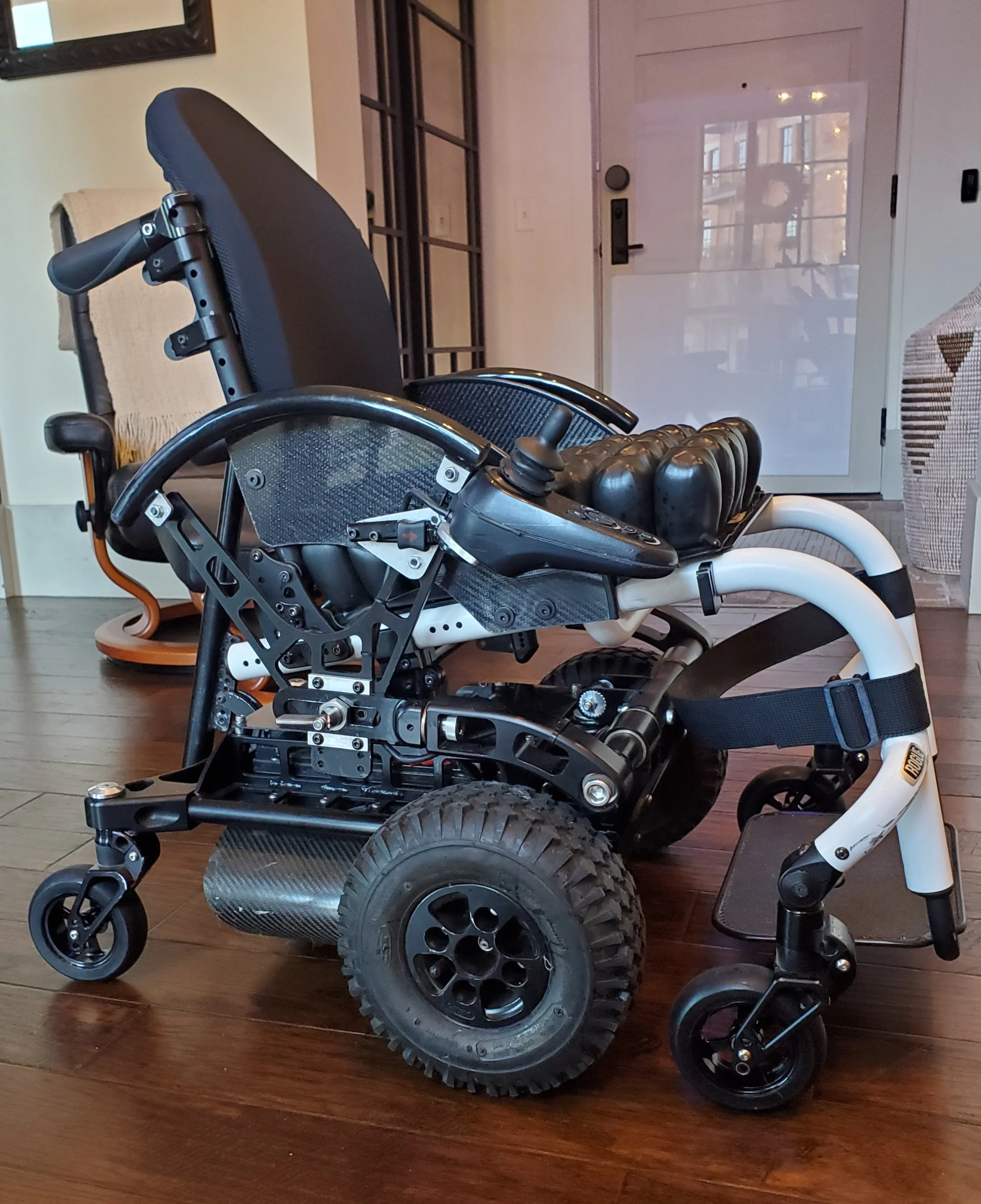A modern wheelchair with large rear wheels, smaller front wheels, and a black cushioned seat, placed on a wooden floor inside a home.