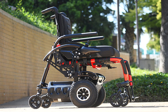 A red Power-Flex wheelchair device outdoors on sidewalk with trees and bushes in the background.