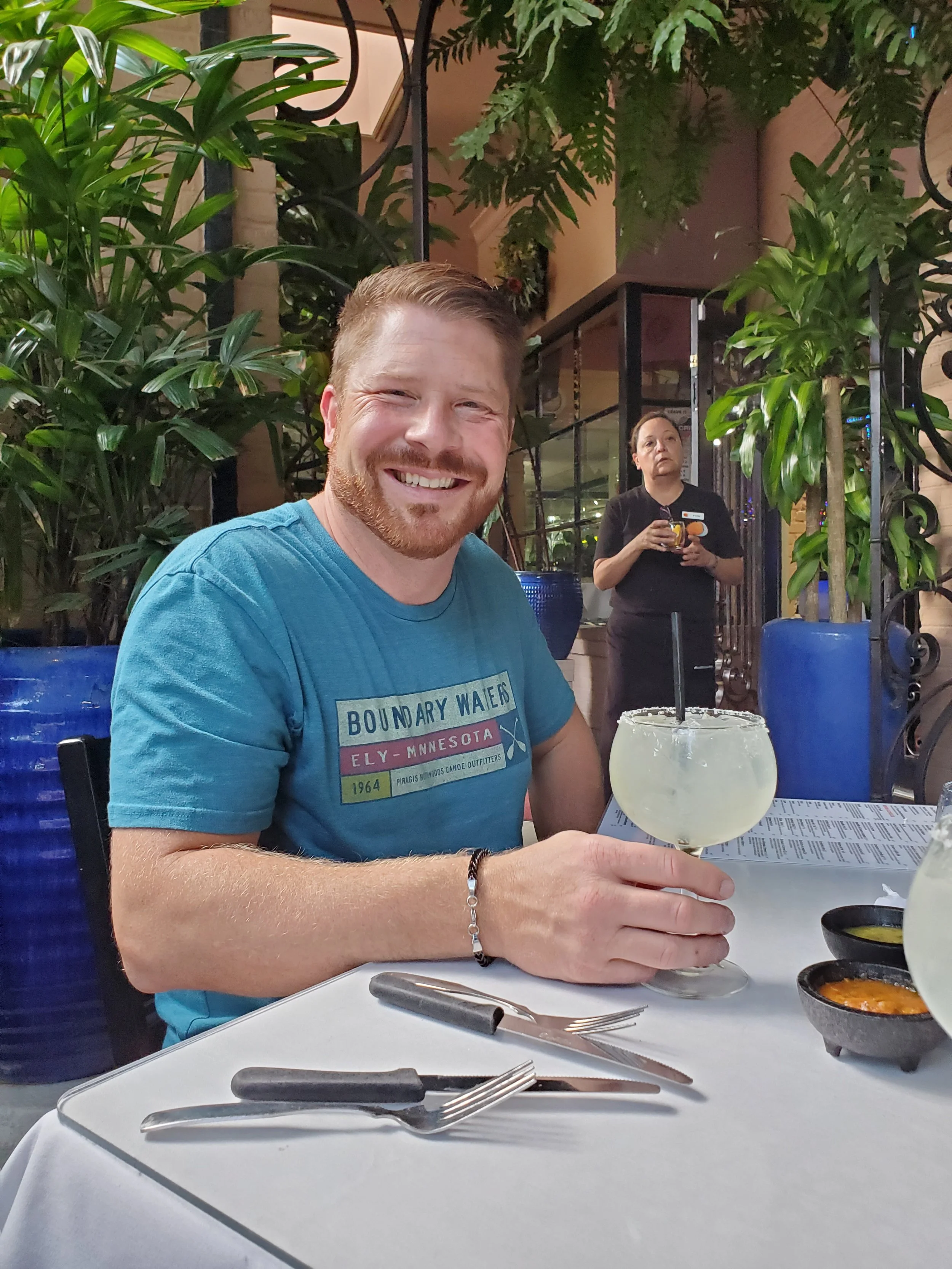 A man with a beard and short hair smiling while holding a large margarita glass in a restaurant setting. He is wearing a blue t-shirt. In the background, a female staff member wearing a black uniform and a name tag is holding a drink. The scene featu