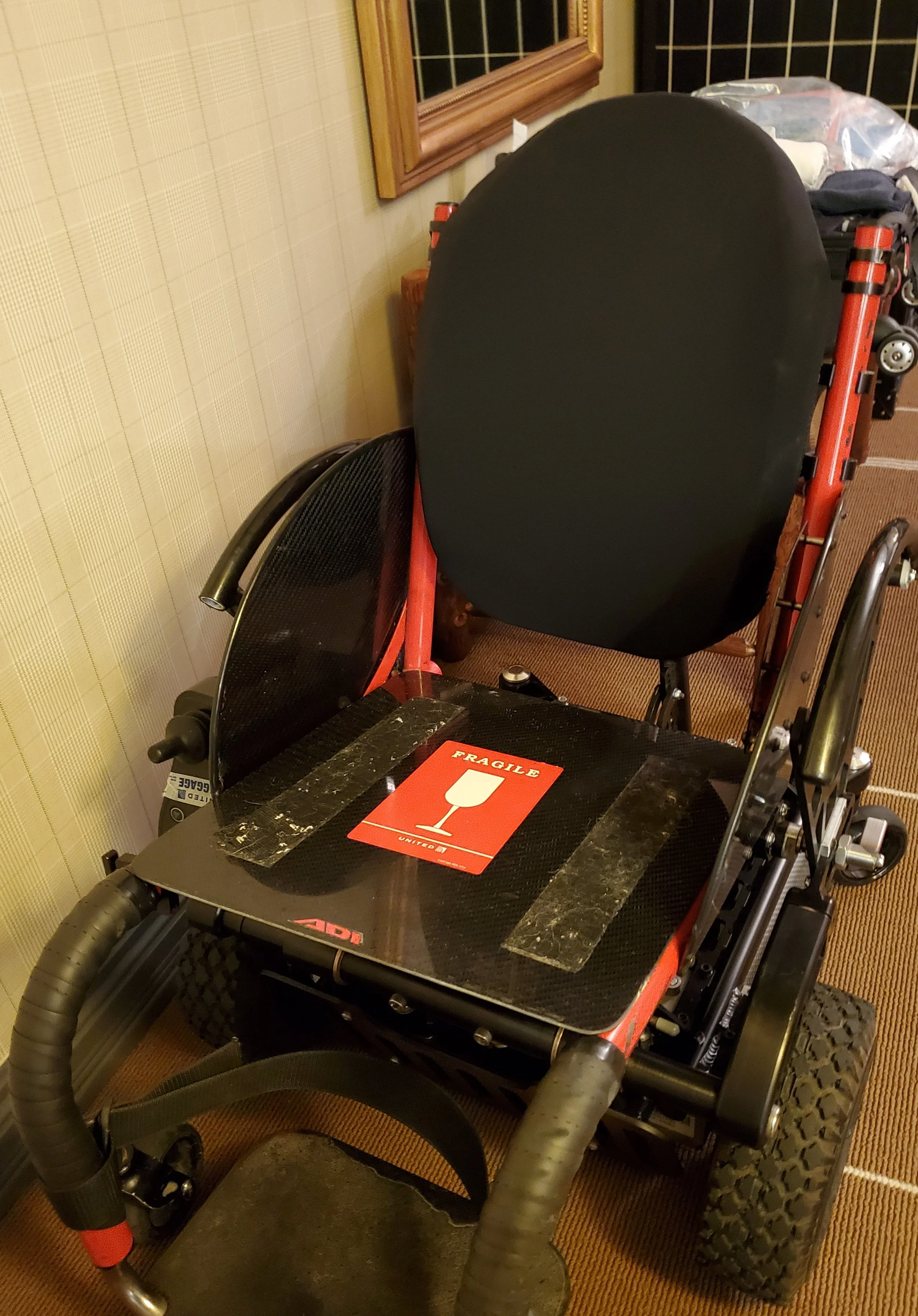 A wheelchair with a red frame and black seat. The wheelchair has a clipboard with a red landmark or fragile sign attached to the seat and is positioned next to a wall with beige patterned wallpaper. There is a plastic bag on a table in the background