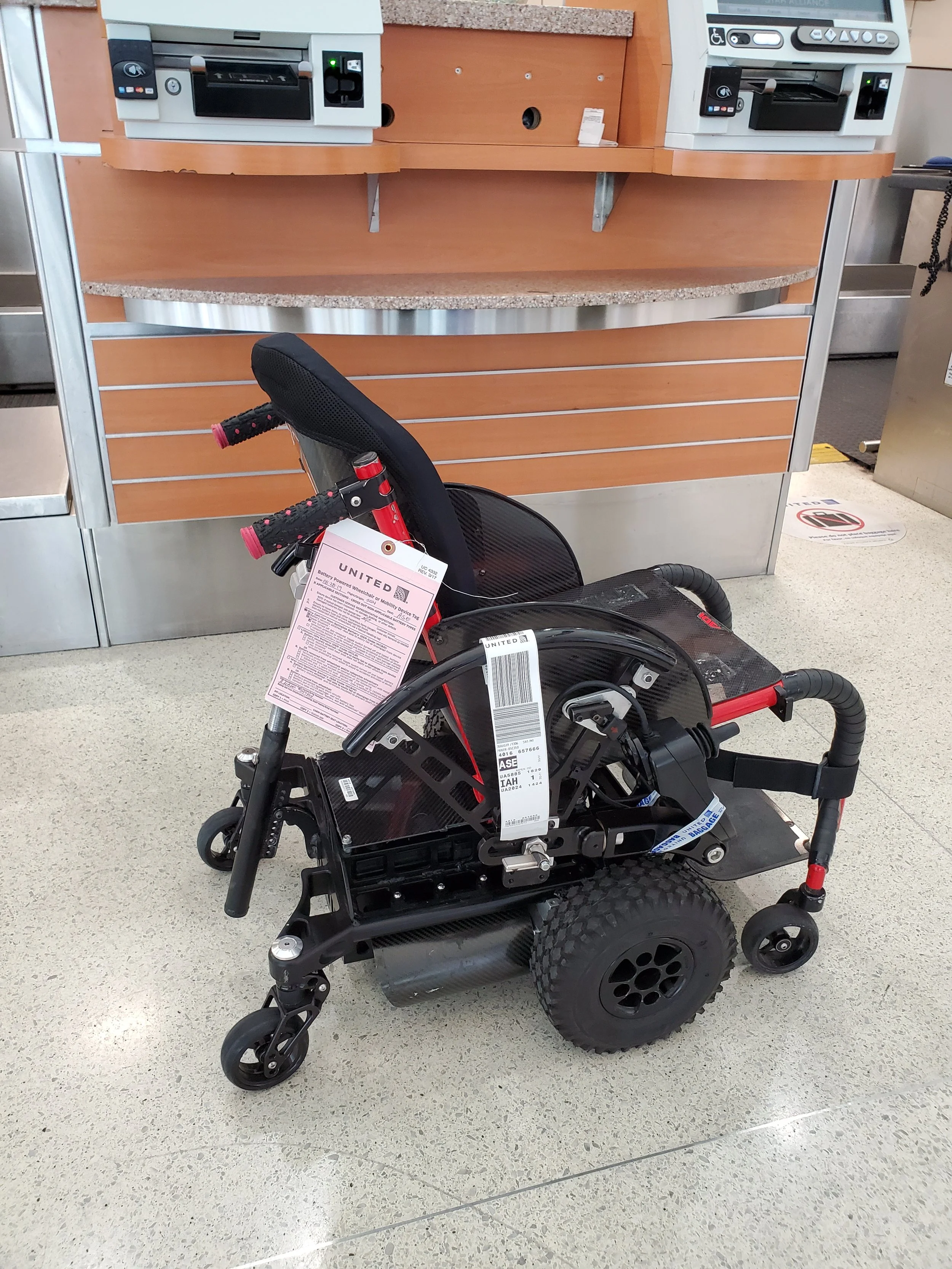 Mobility scooter with warning tags on the seat and frame, standing in front of check-in kiosks in an airport terminal.