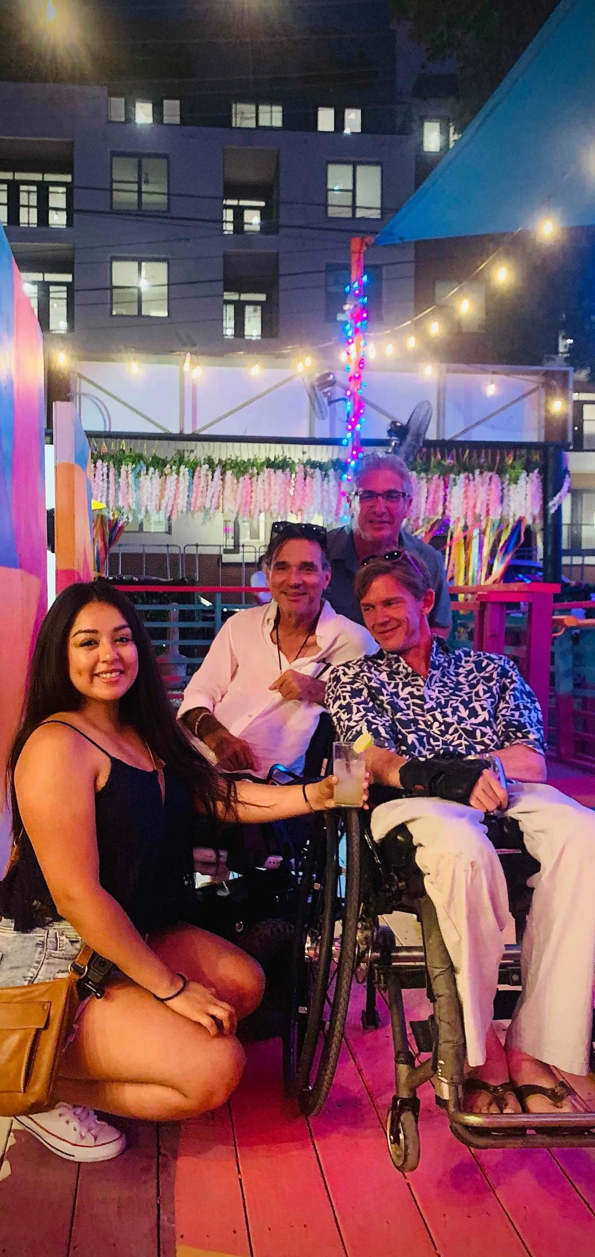 Four people at a party or celebration, with colorful lights and decorations in the background. One woman is kneeling in the front, holding a drink, while three men are sitting or standing behind her, smiling.
