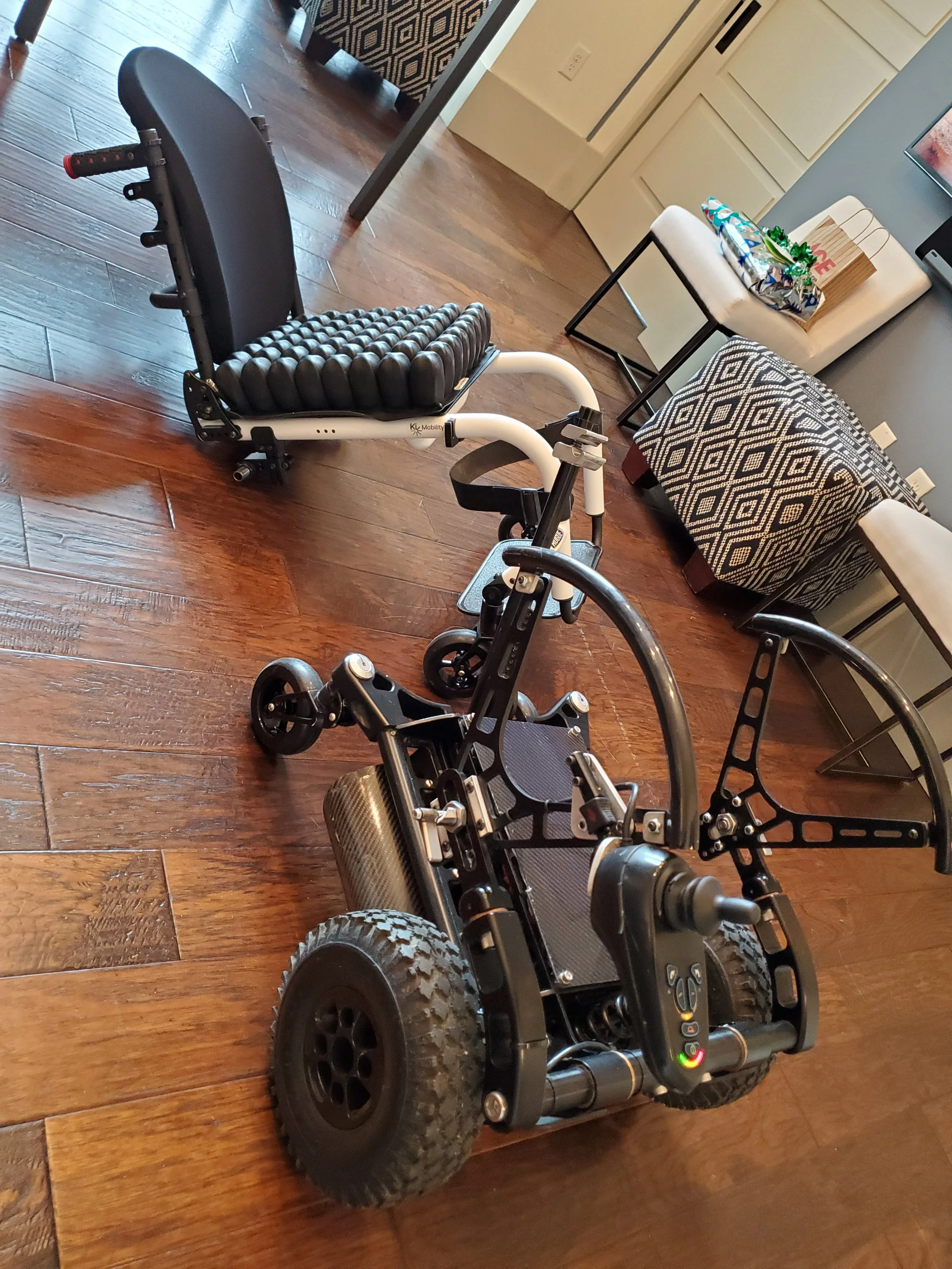 A black power wheelchair with large tires and a padded seat, positioned on a hardwood floor inside a home, near a beige chair with gifts on top.