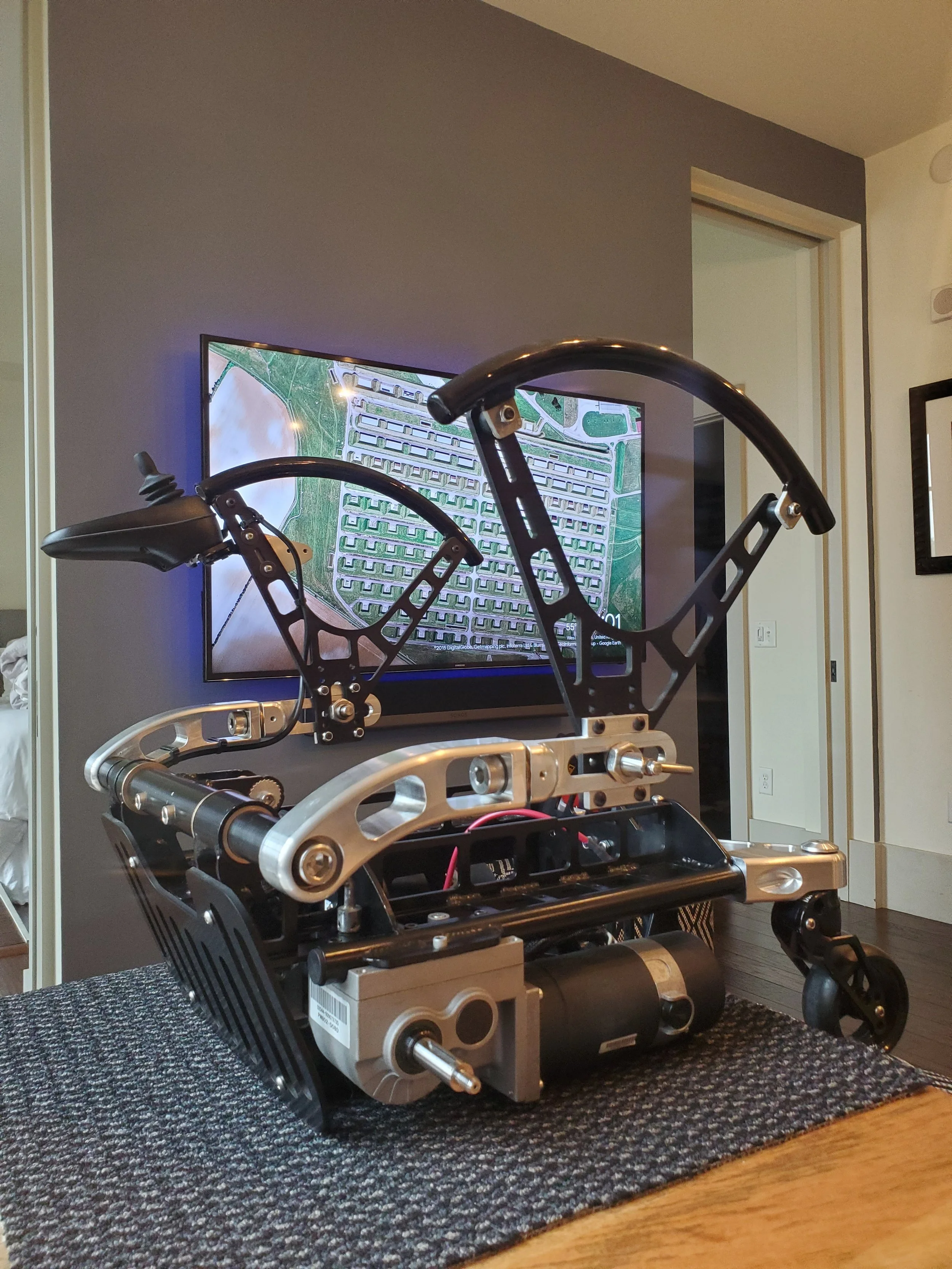 Unassembled electric wheelchair with a metal frame, mounted on a black textured mat, positioned in front of a gray wall and TV screen in a room.