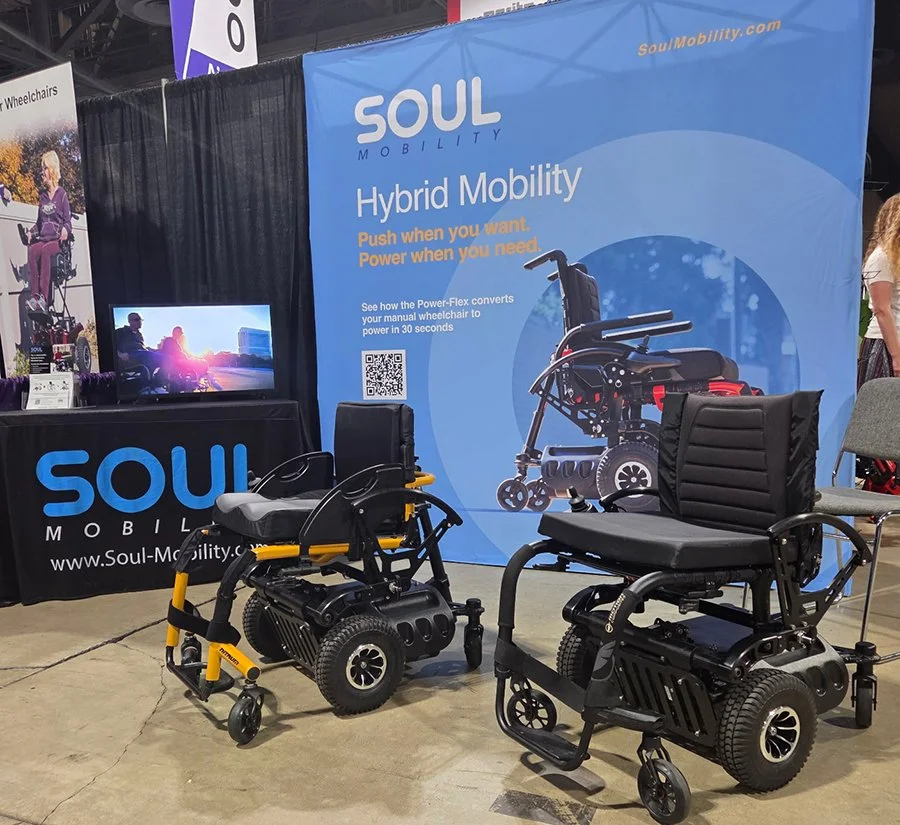 Two power wheelchairs displayed at a Soul Mobility booth with a large blue banner promoting hybrid mobility and controls. A small TV screen shows a person using a wheelchair outdoors.