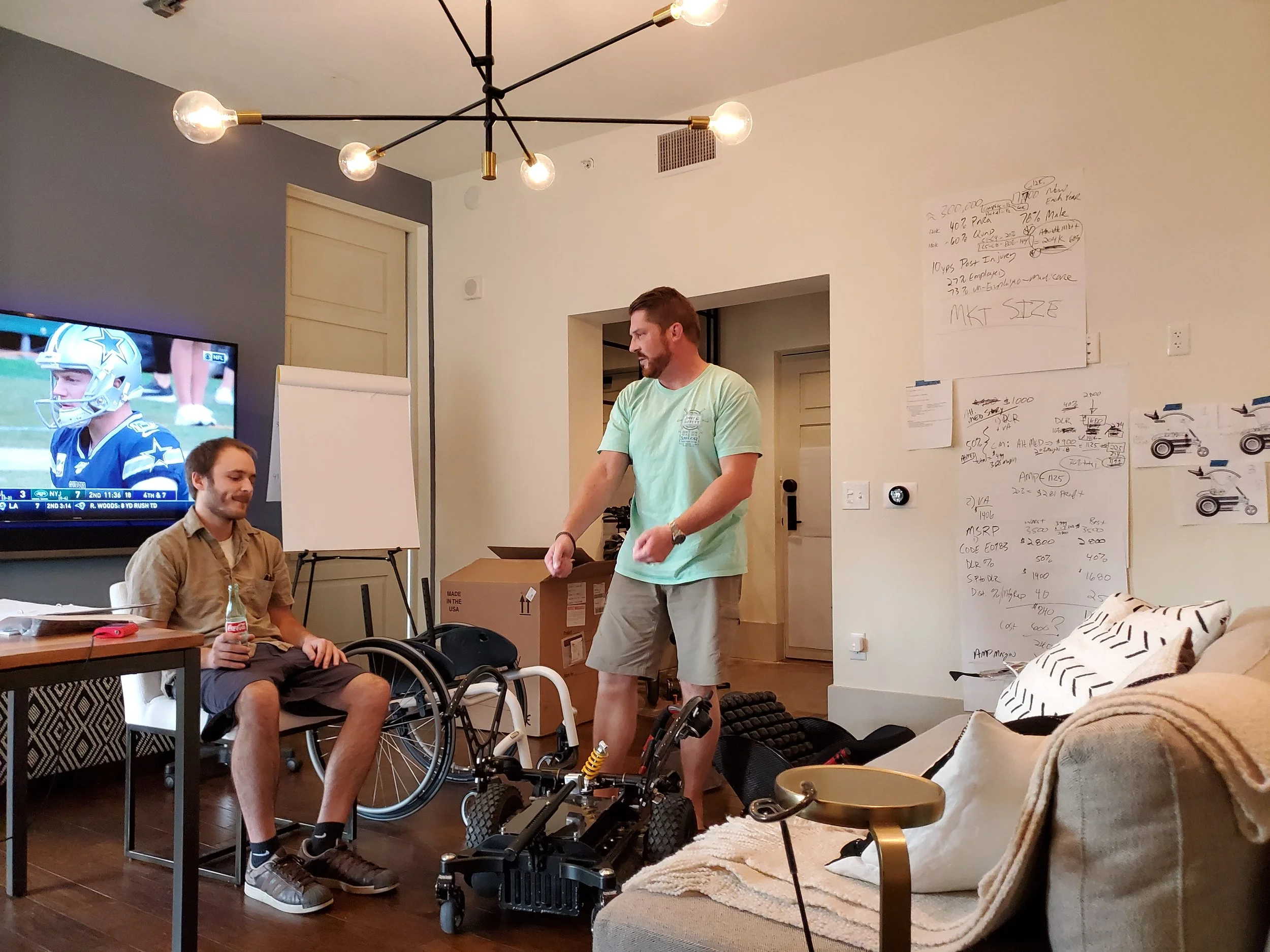Three men are in a living room, one is seated in a wheelchair holding a water bottle, another is standing holding a bicycle, and the third man is partially visible in the foreground. The living room has a television, a whiteboard with notes, and vari