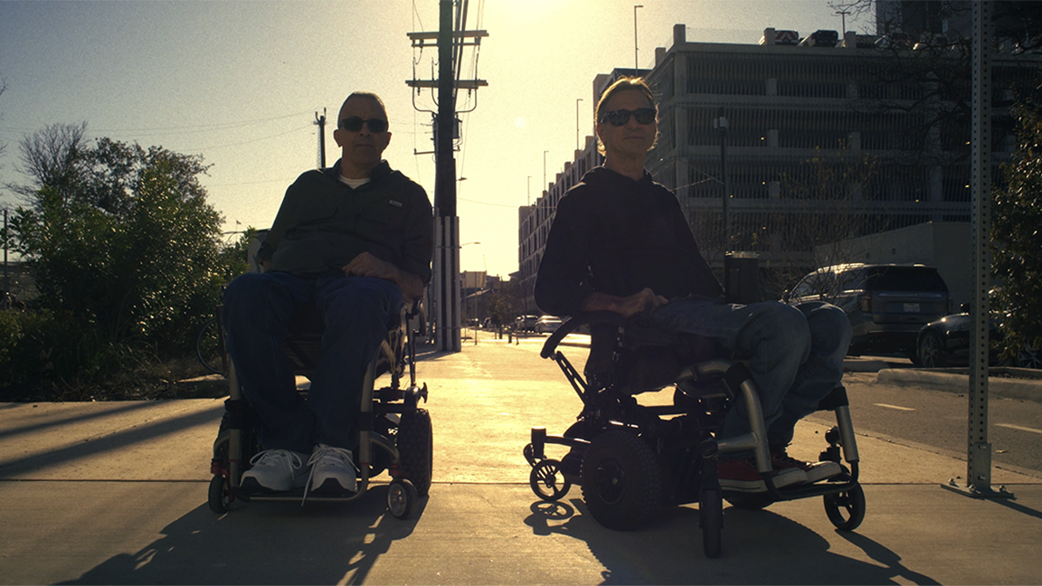 Two people sitting in wheelchairs on a sidewalk with sunlight behind them, in an urban setting with buildings and parked cars.