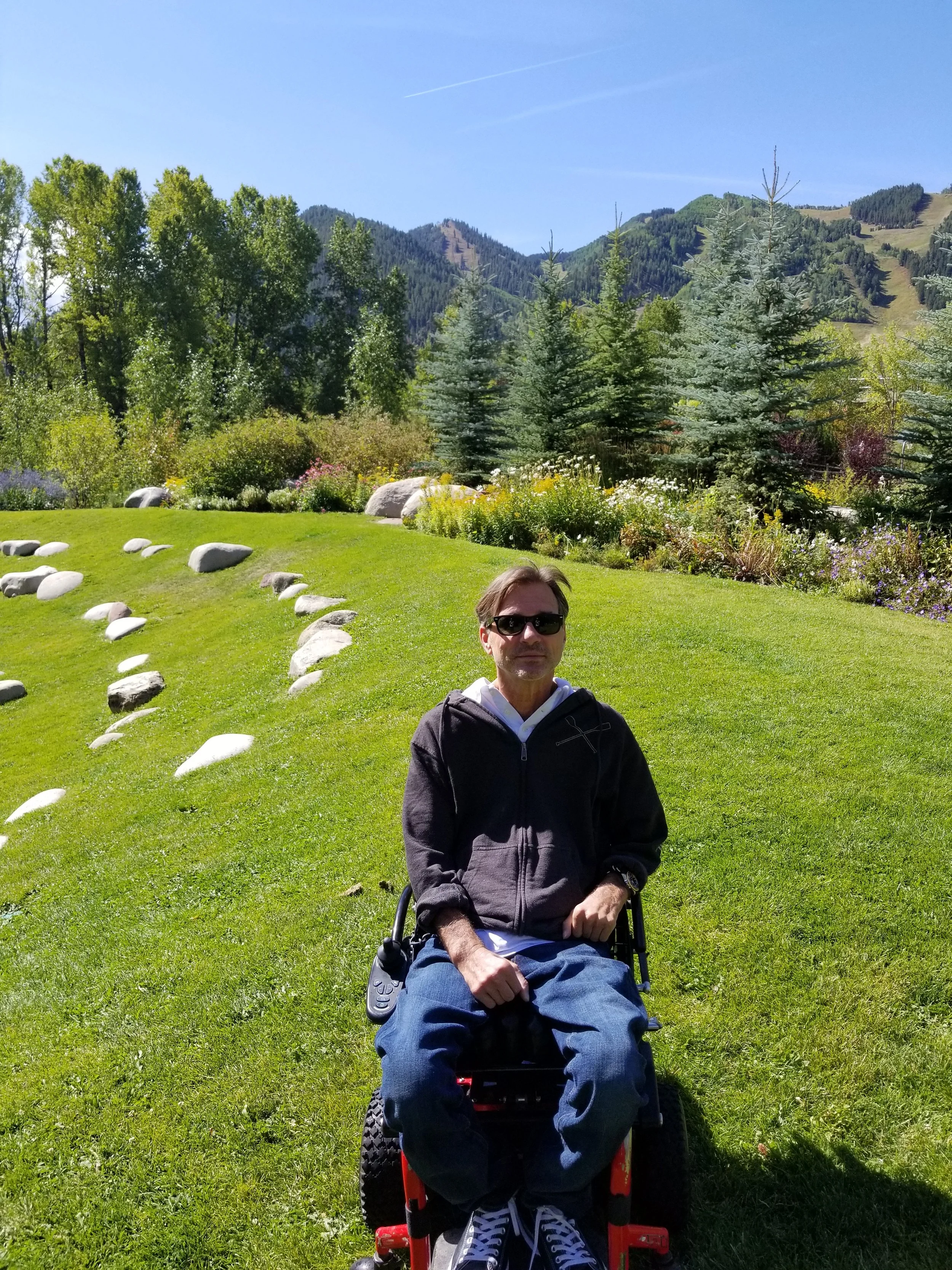 A man in a wheelchair with sunglasses, sitting outdoors on a grassy area with a mountain landscape in the background, including trees and clear blue sky.