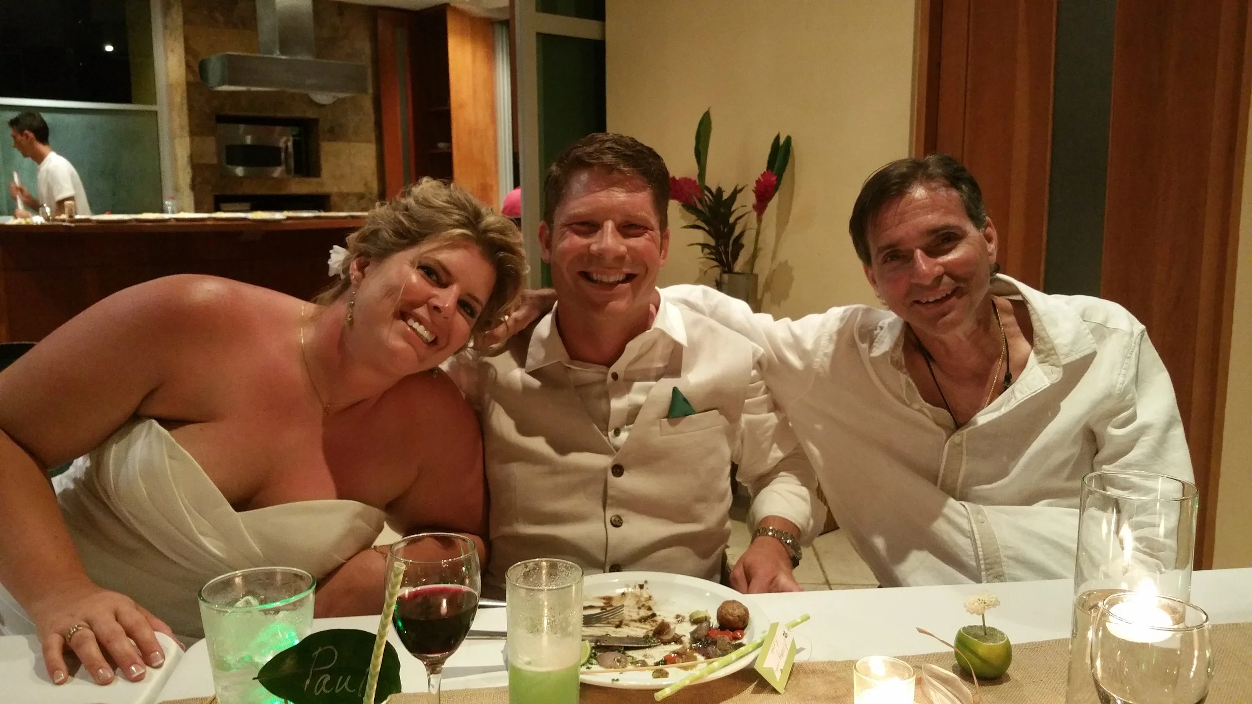 Three people sitting at a dinner table, smiling for the camera, with glasses of drinks and a partially eaten meal in front of them, in a warmly lit indoor setting.