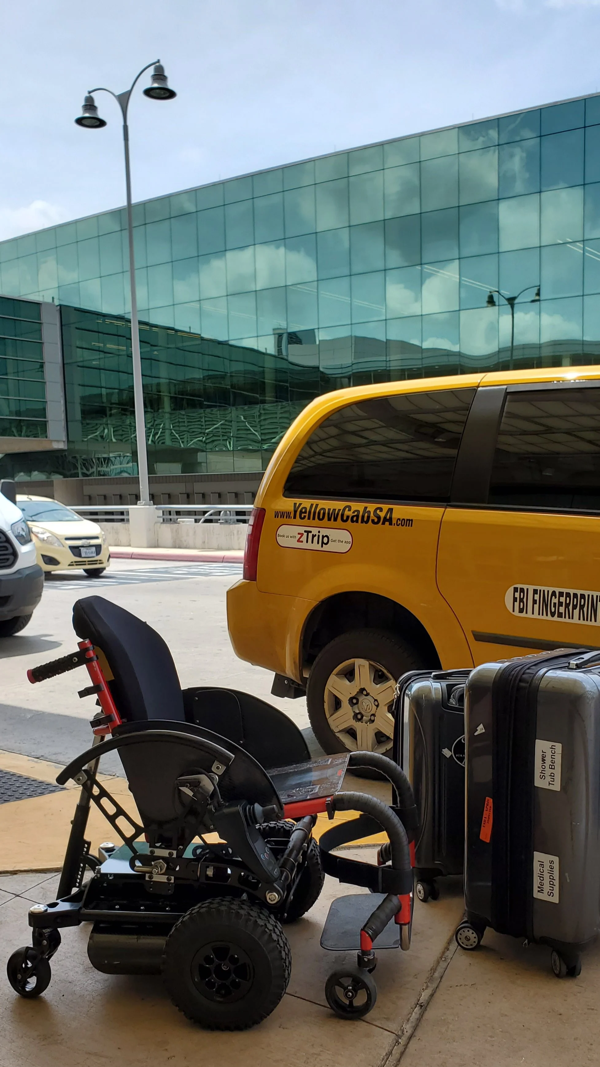 A yellow cab with the website YellowCabSA.com parked near a modern glass building. In the foreground, there is a wheelchair, a suitcase labeled Medical Supplies, and luggage, all near a sidewalk with a bench and a bicycle.