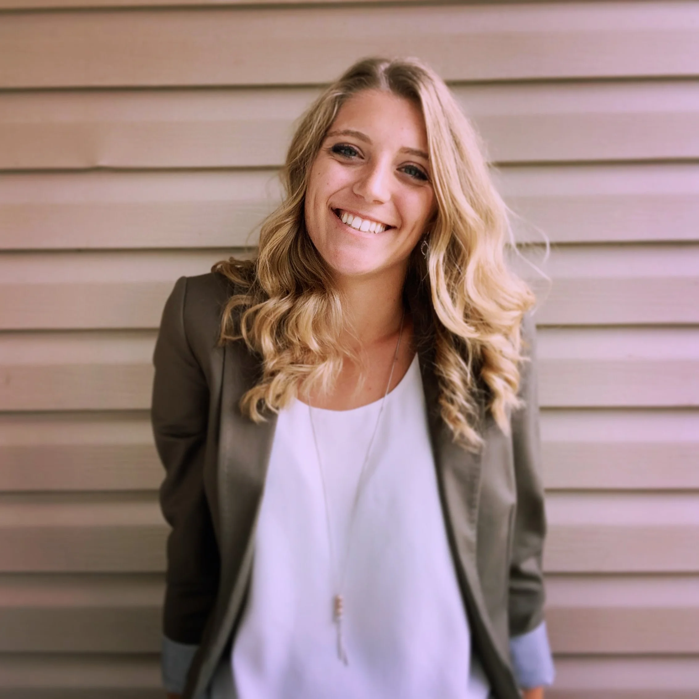 A woman with long blonde curly hair and a wide smile, standing in front of beige horizontal siding.