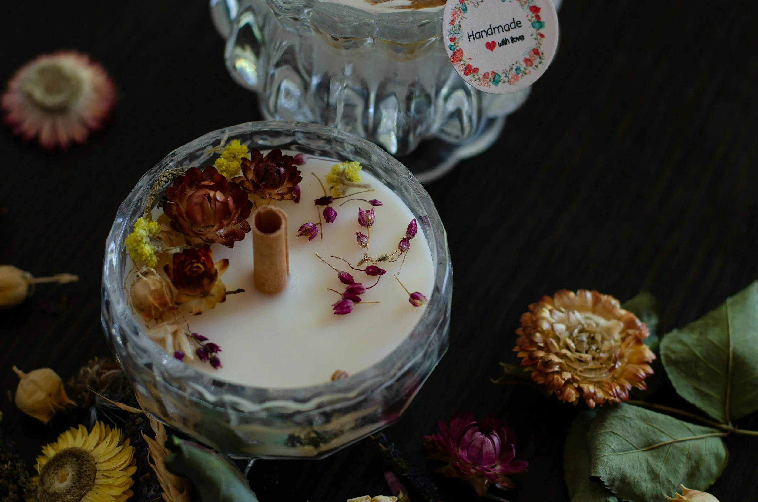 Close-up of a white candle in a glass holder decorated with dried flowers and rosebuds, surrounded by scattered dried flowers and leaves on a dark surface. A tag on a glass adorns the candle, reading "Handmade with love" with floral design.