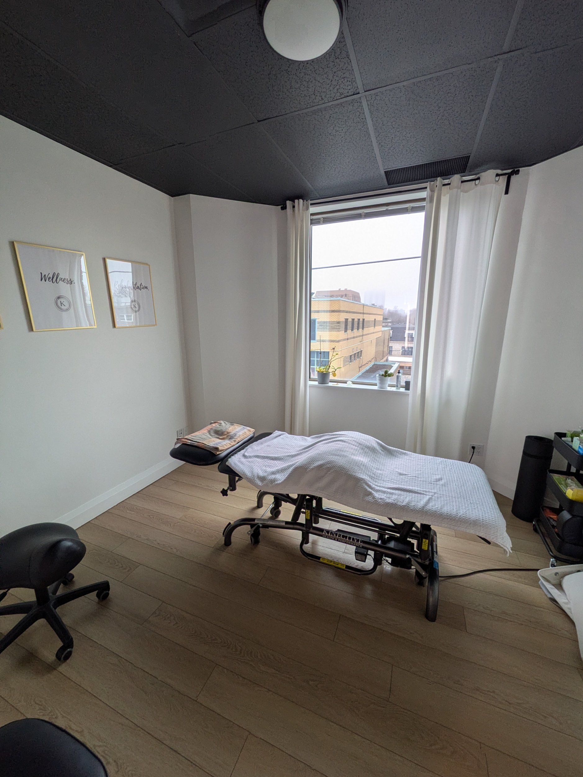 A room with a therapy table covered with a white sheet, a small pillow, and a folded blanket. There is a black office chair with wheels and a puzzle piece rug. The room has white walls with framed prints reading 'Wellness' and 'Relaxation,' a window with white curtains, and a view of buildings outside.