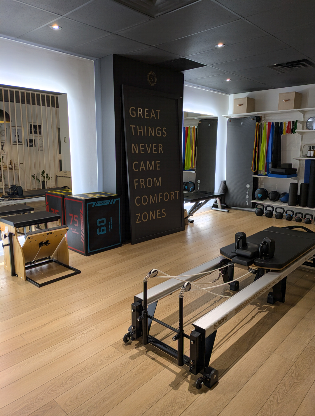 A fitness studio with workout equipment including a reformer machine, plyometric boxes, kettlebells, medicine balls, and resistance bands. A large motivational sign reads, "Great things never came from comfort zones."