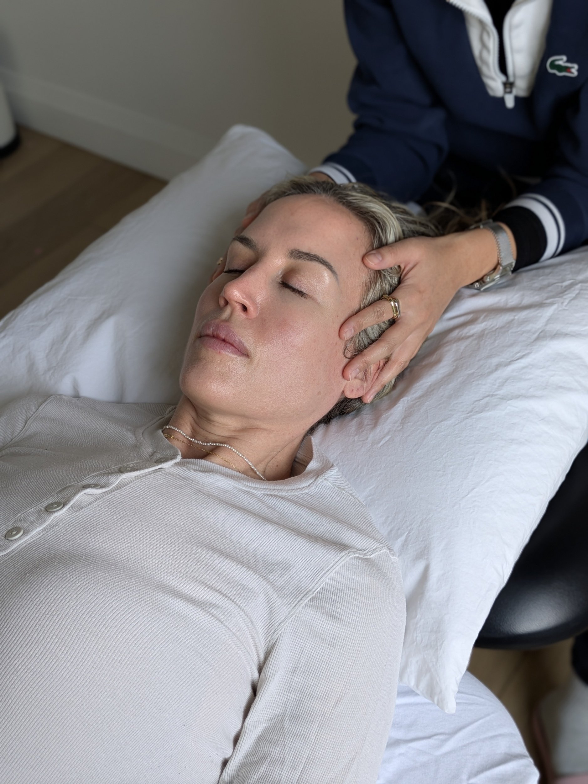 A woman with short blonde hair lying on a bed with her eyes closed, receiving a head massage from a person wearing a dark blue jacket with white accents.