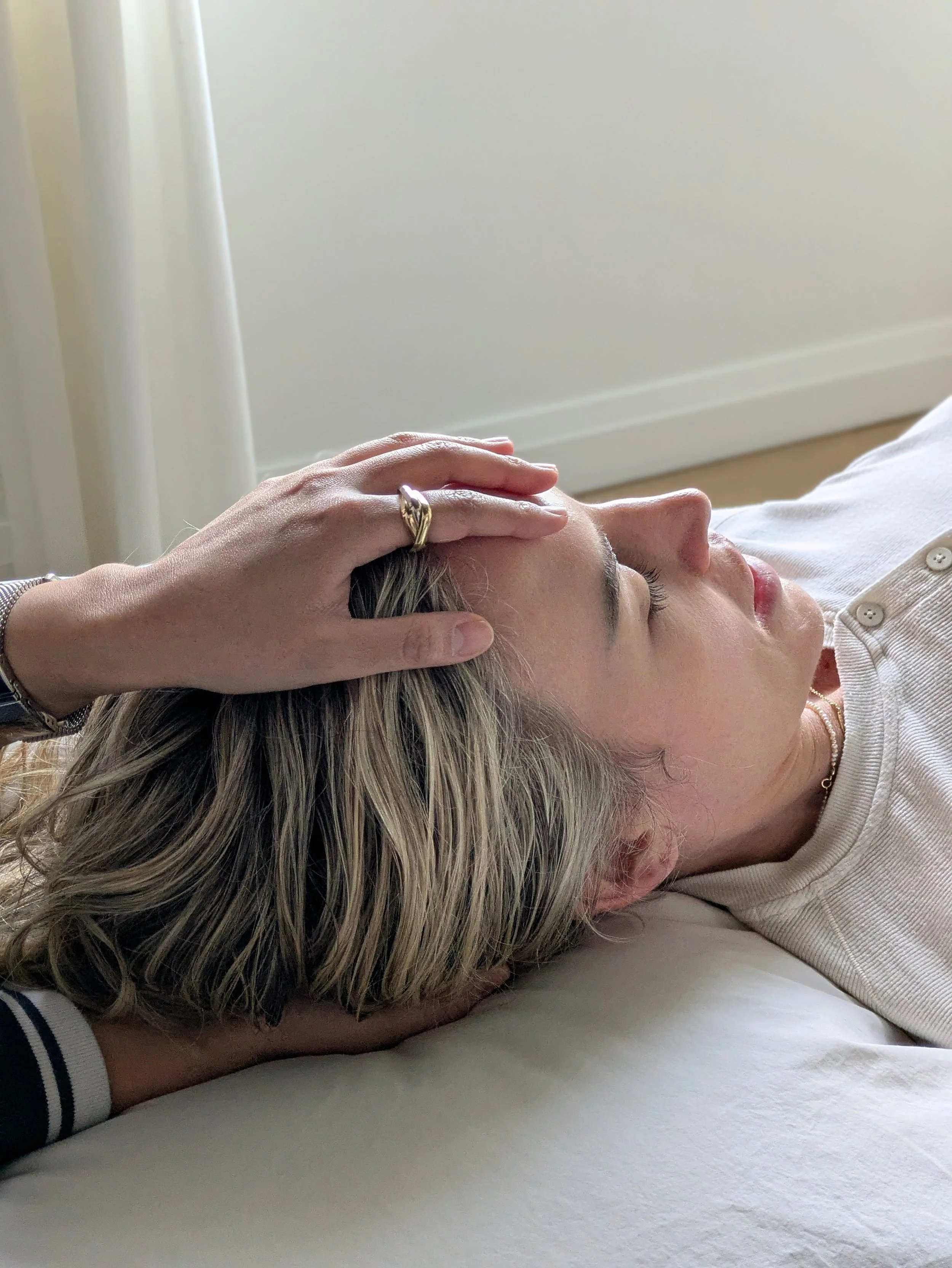 A woman with blonde hair lies on her side on a bed with white sheets, resting her head on a pillow. A hand with a ring rests on her forehead. She is wearing a light-colored top with buttons.