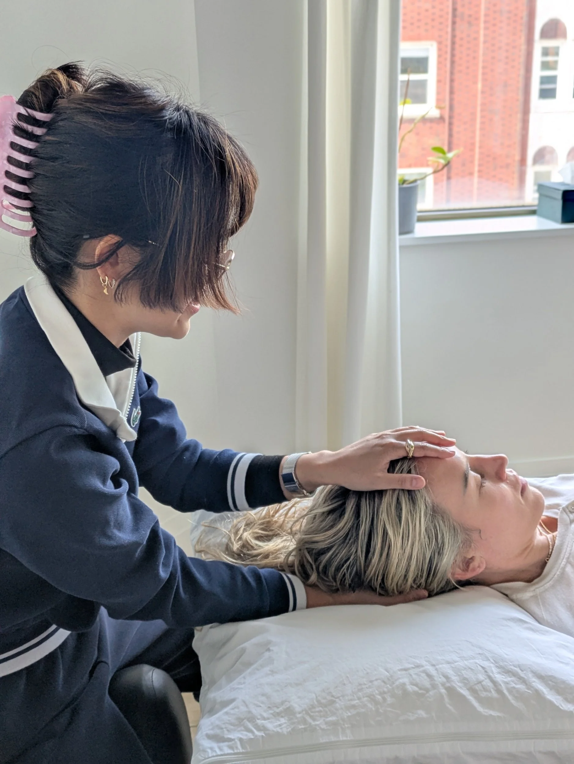 A woman with short brown hair and earrings gives a head massage or treatment to another woman with blonde hair lying on a bed, in a room with a window and curtains.