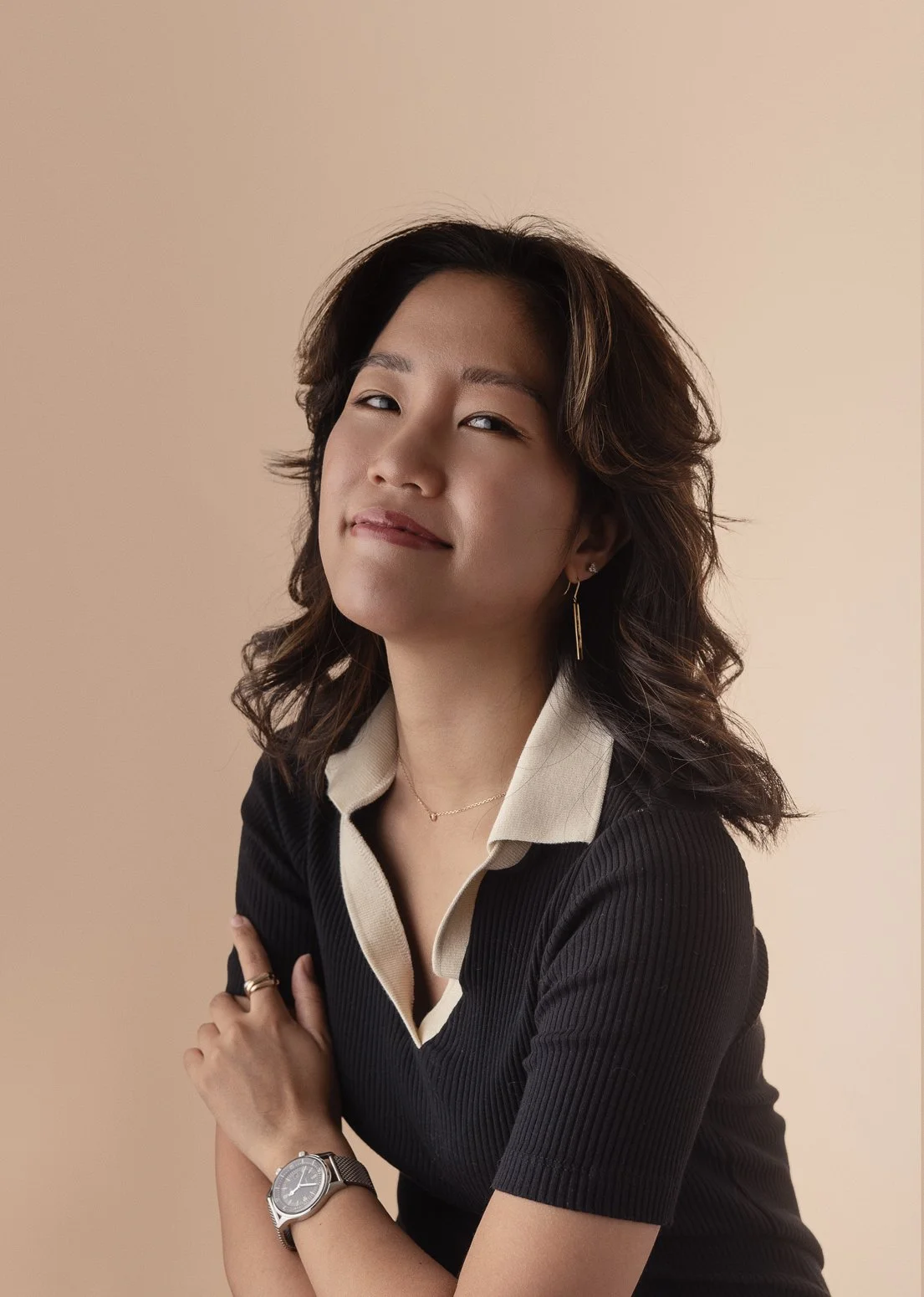 A woman with wavy dark hair wearing a black top with a beige collar, accessorized with a watch, rings, a necklace, and a earring, posing with her arms crossed against a beige background.