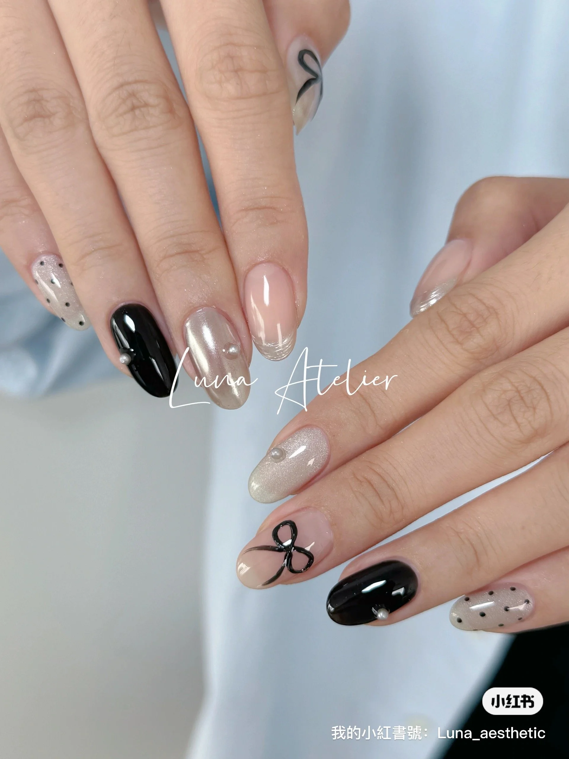 Close-up of a woman's hands showcasing manicured nails with various designs including black, nude, pearl, and polka dot patterns, some with decorative accents like small pearls and a bow, against a light background.
