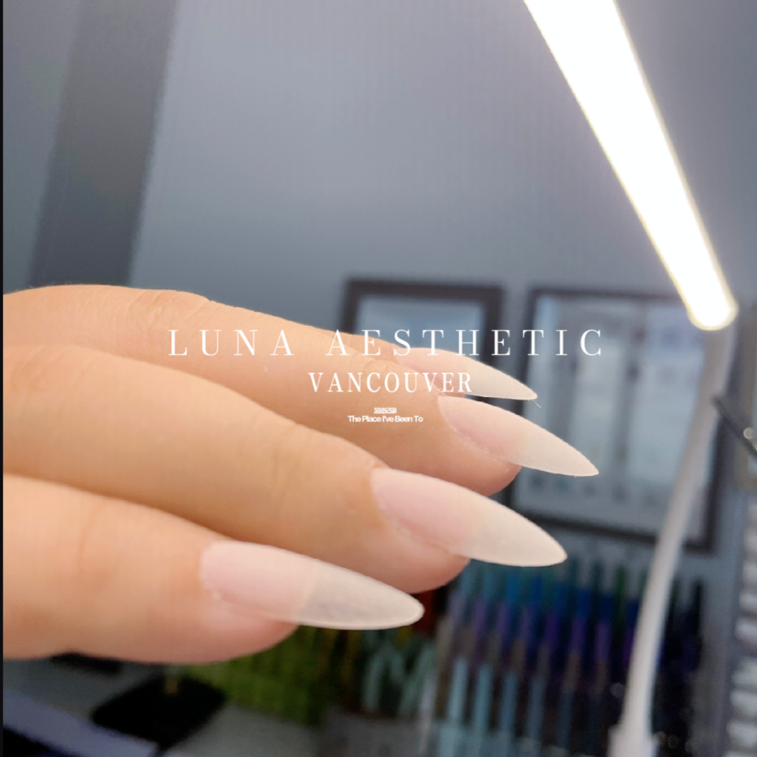 Close-up of a hand with long, pointed, nude-colored acrylic nails, with text overlay reading 'LUNA AESTHETIC VANCOUVER'.
