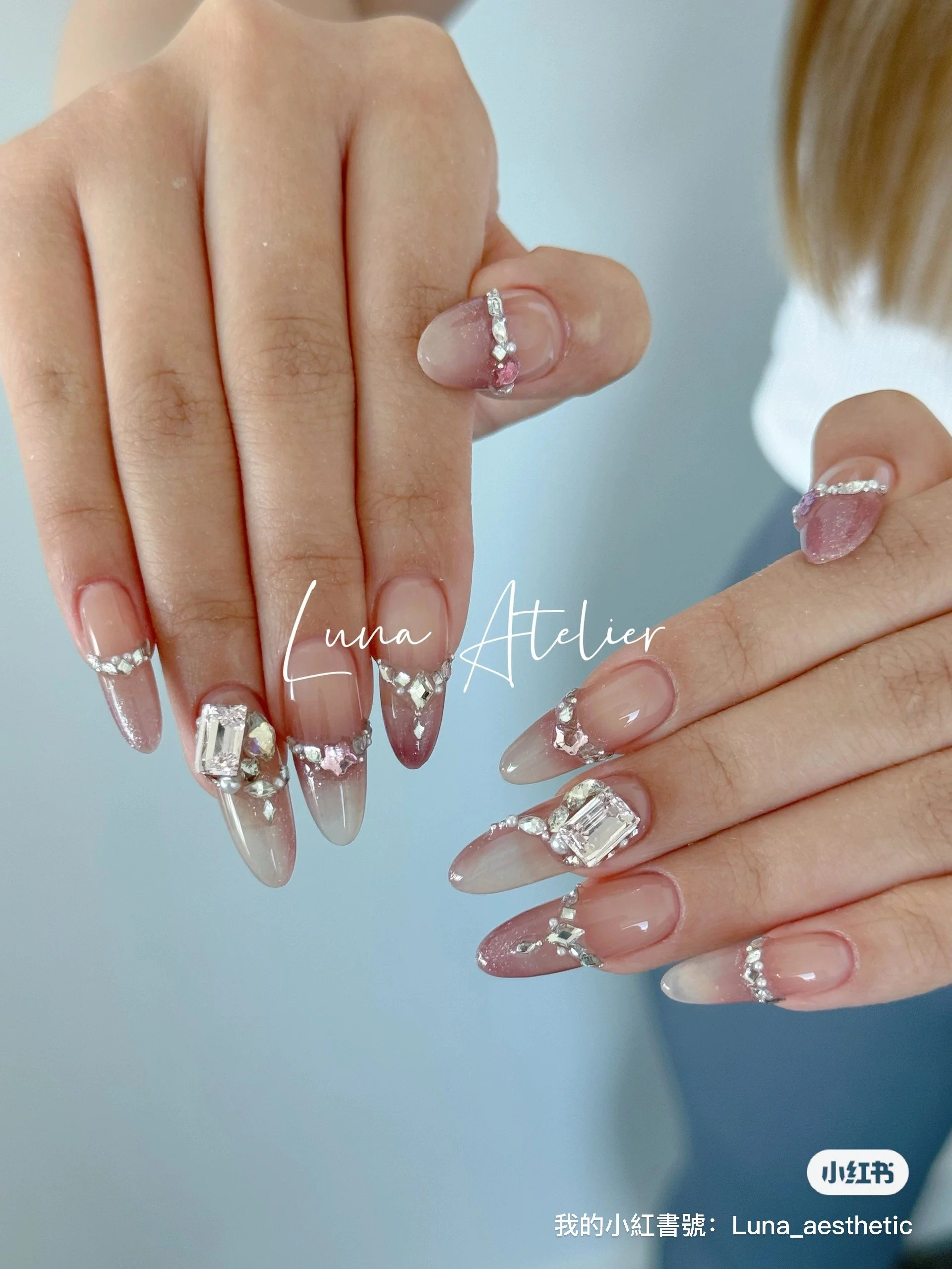 Close-up of hands with long, almond-shaped nails adorned with rhinestone jewelry and pink accents.